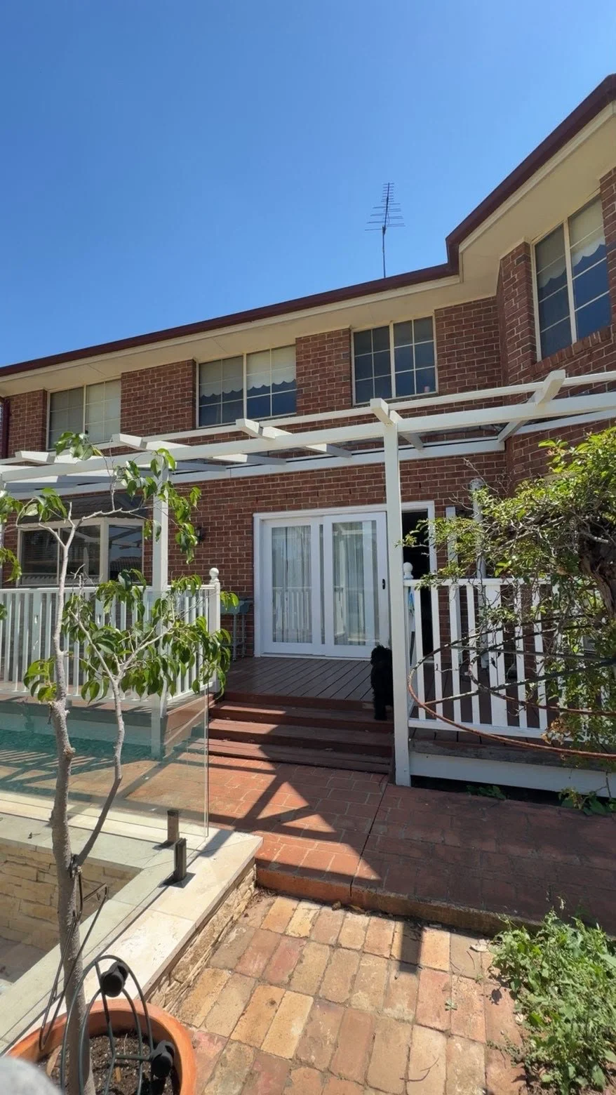 Brick home with white pergola, glass doors, deck, garden, and swimming pool under a clear blue sky