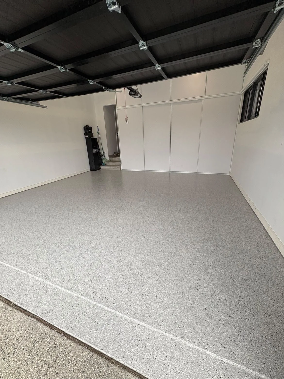 Empty residential garage with a speckled gray epoxy floor, open overhead door, white walls, storage cabinet, and small window