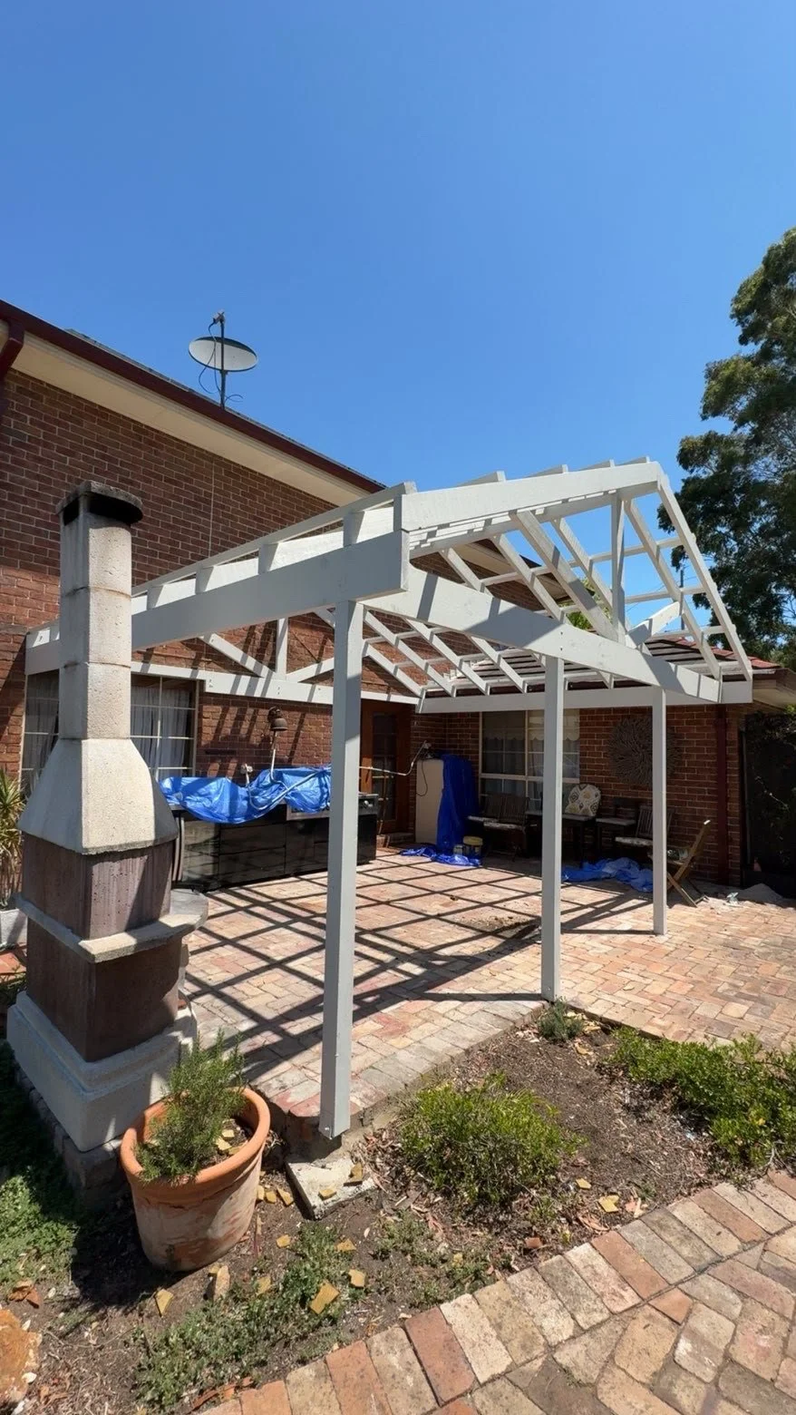 White timber patio pergola under construction beside a brick house, with outdoor fireplace, satellite dish, brick paving, and potted plants