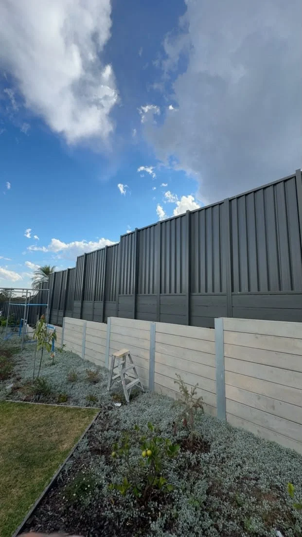 Gray corrugated metal privacy fence above a concrete block wall beside a landscaped garden under a partly cloudy blue sky