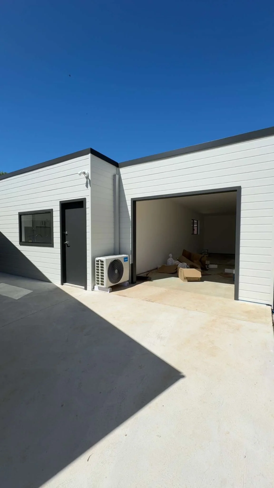 Modern white-sided detached garage with an open bay, black door, window, and outdoor HVAC unit under a clear blue sky