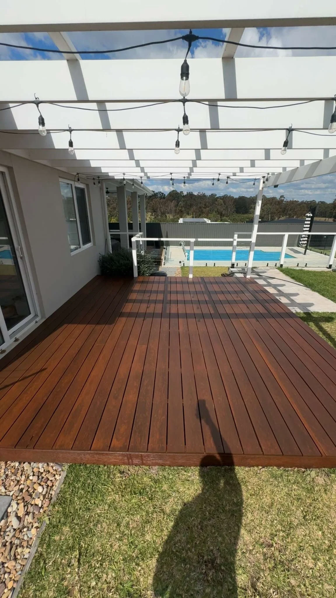 Modern backyard with a covered wooden deck, string lights, white railings, and a swimming pool