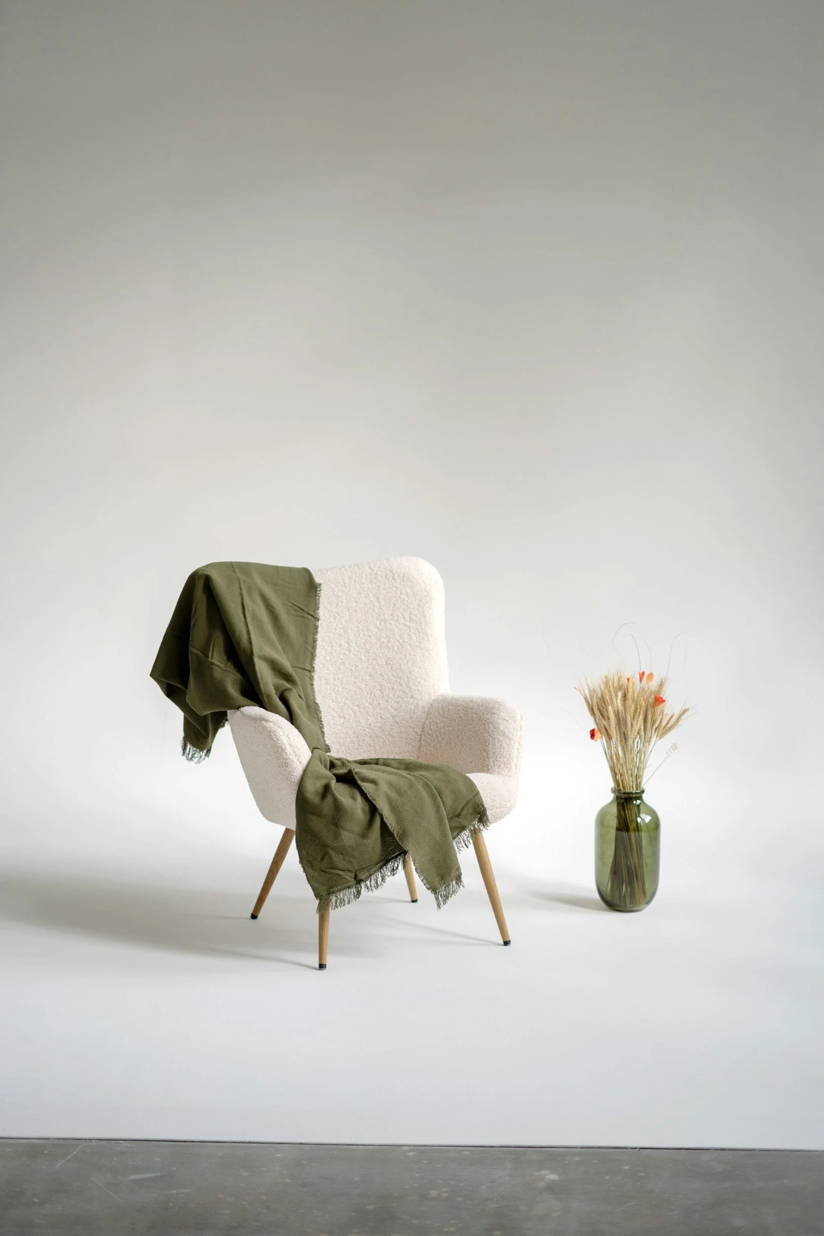Chaise blanche en tissu avec un vêtement vert dessus, vase avec des plantes séchées à côté, mur blanc en fond.