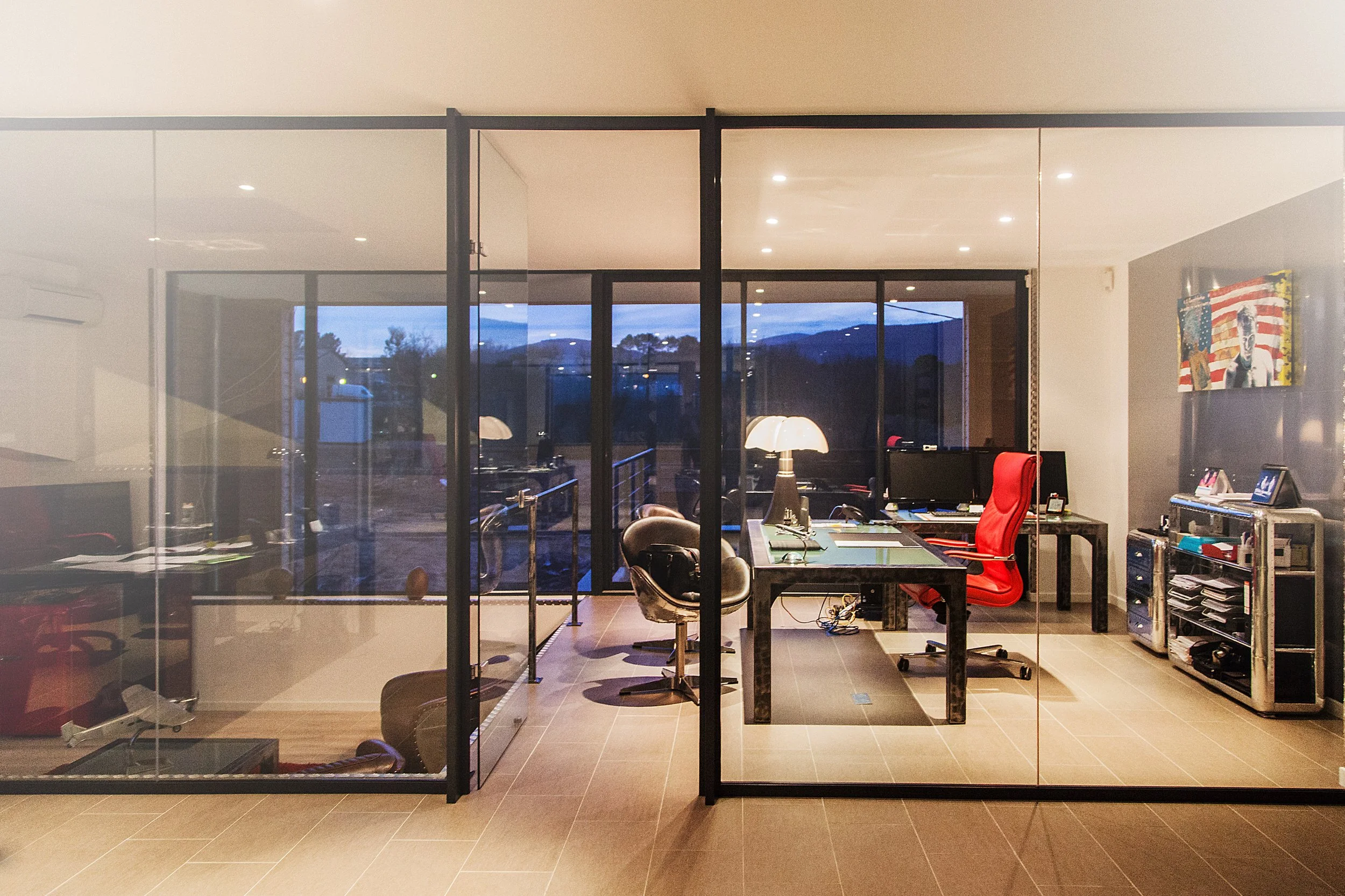 Bureau moderne avec table de travail, écran, lampes et fauteuils, vue à travers une grande fenêtre avec paysages et montagnes en arrière-plan.