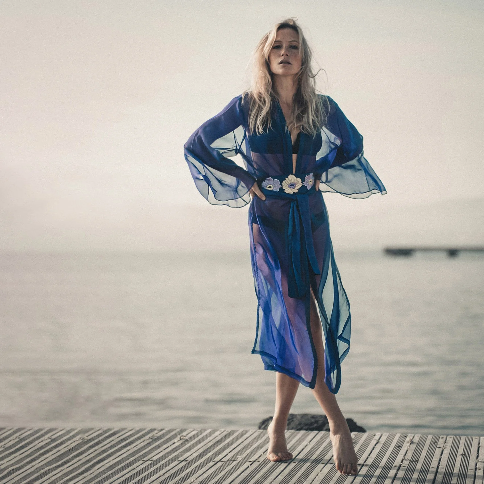 Femme portant une robe en tulle transparente bleue avec des broderies de fleurs blanches, posant sur un quai en bois au bord de l'eau pendant le coucher du soleil.