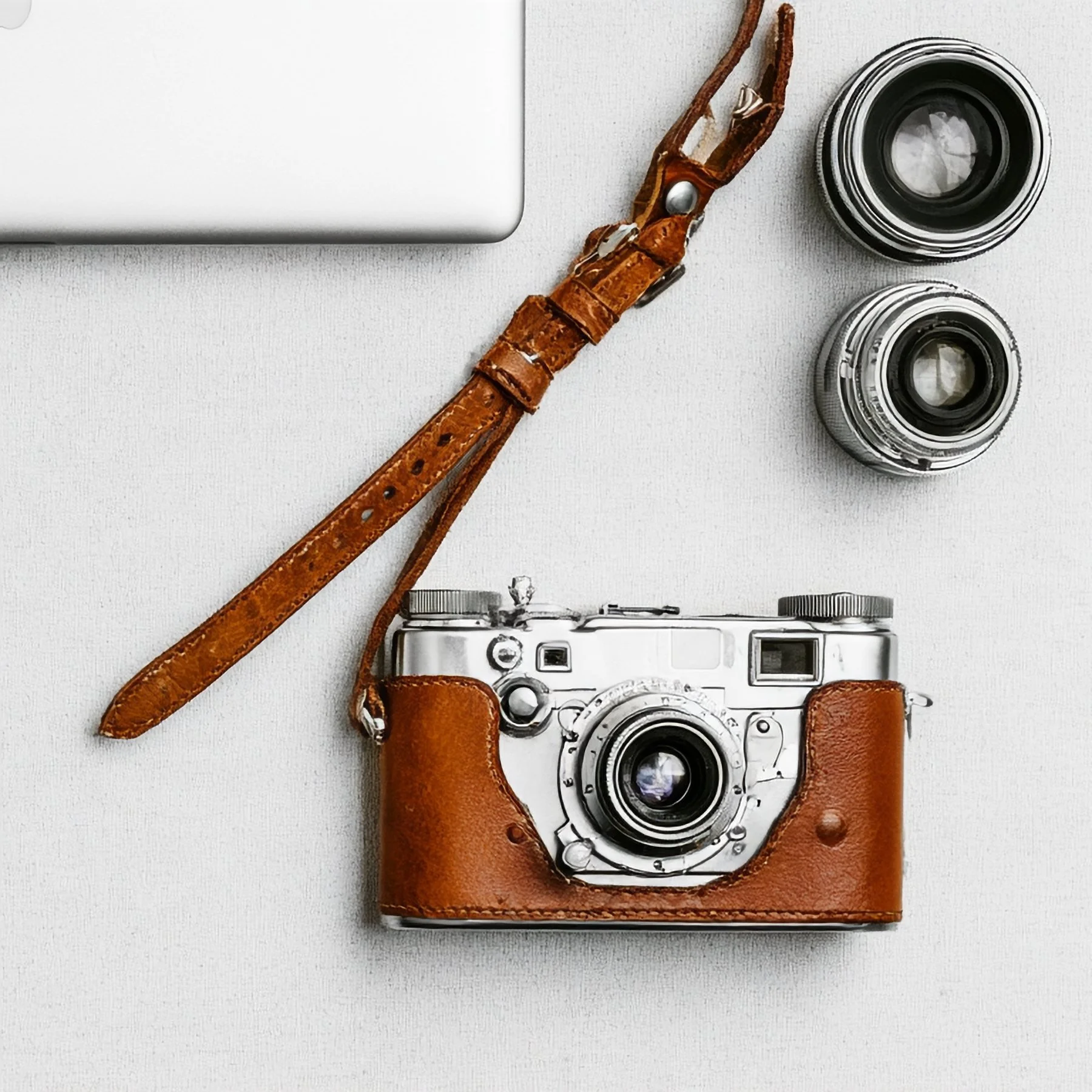 Appareil photo vintage posé dans une étui en cuir marron, avec un objectif supplémentaire, placé sur une surface blanche avec un ordinateur portable visible dans le coin supérieur gauche.