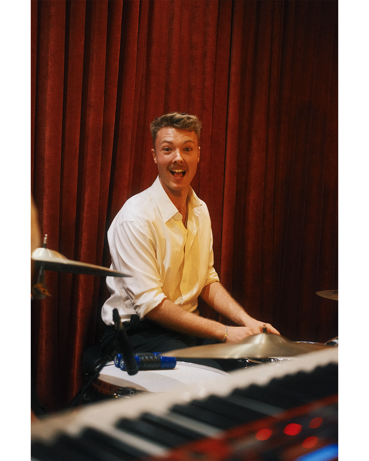A man in a yellow shirt sitting at a keyboard, smiling and looking excited, with a red curtain in the background.