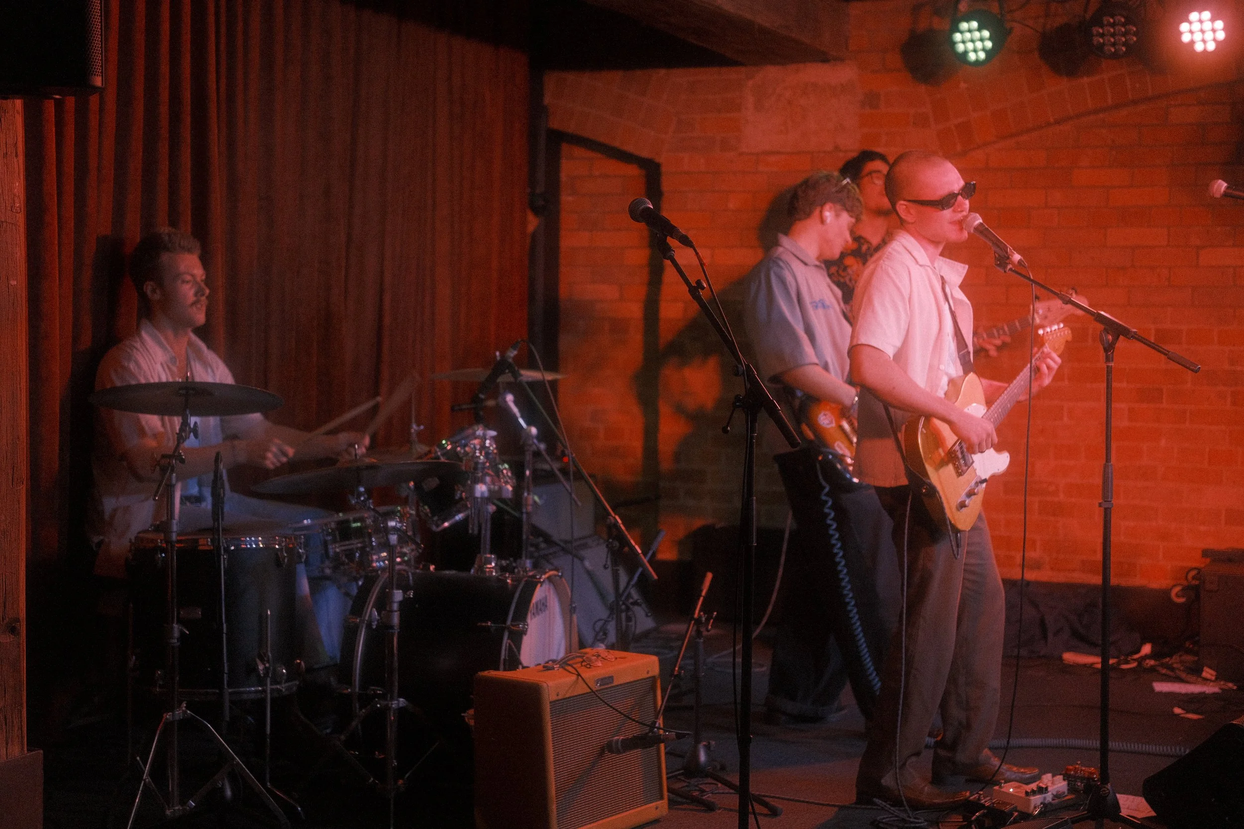 Band performing live on stage in a brick-walled venue, with three musicians visible: a drummer, and two guitarists, one singing into a microphone and wearing sunglasses.