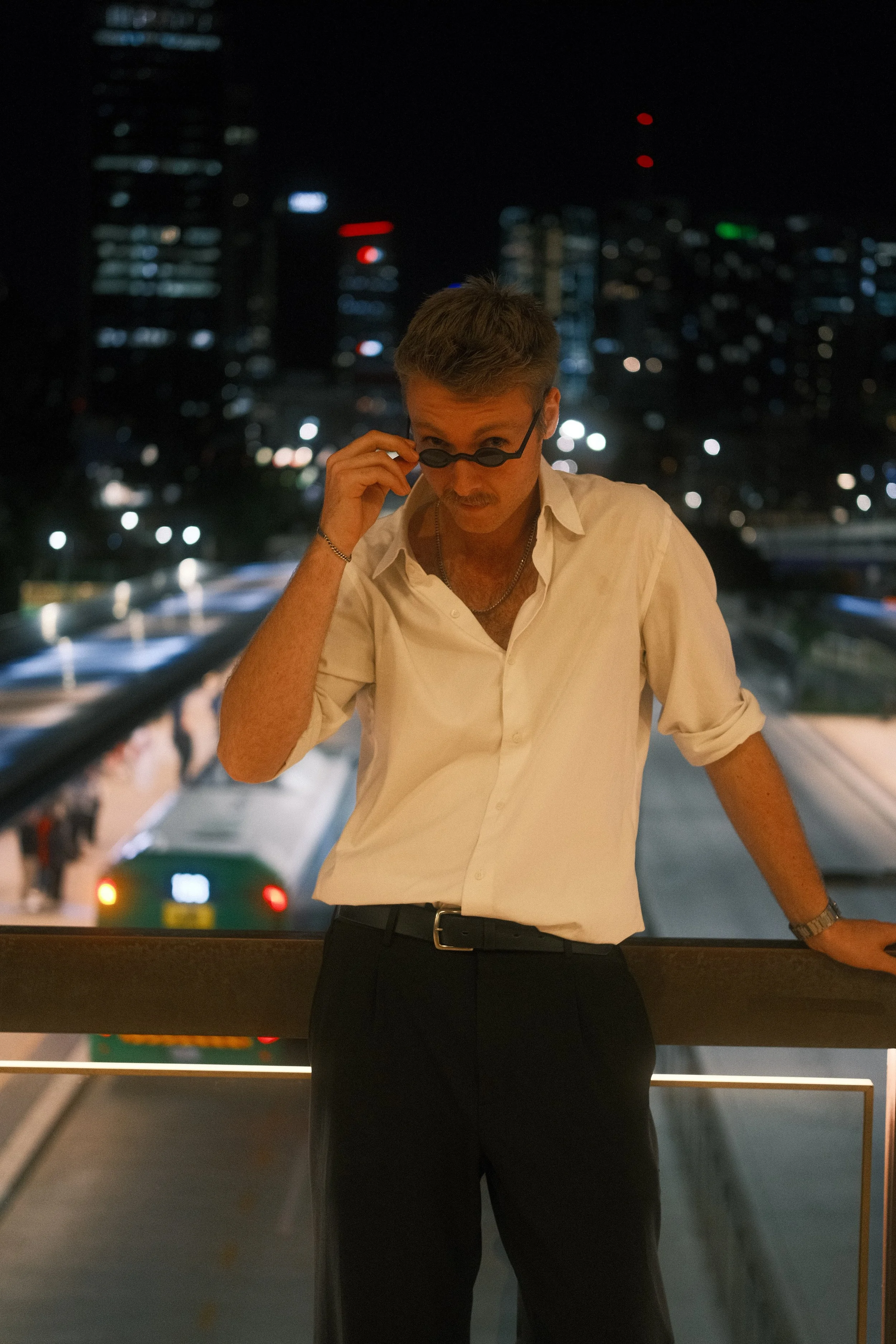A man in a white shirt and black pants standing on a bridge at night, wearing sunglasses and leaning against the railing with city skyscrapers and a bus in the background.