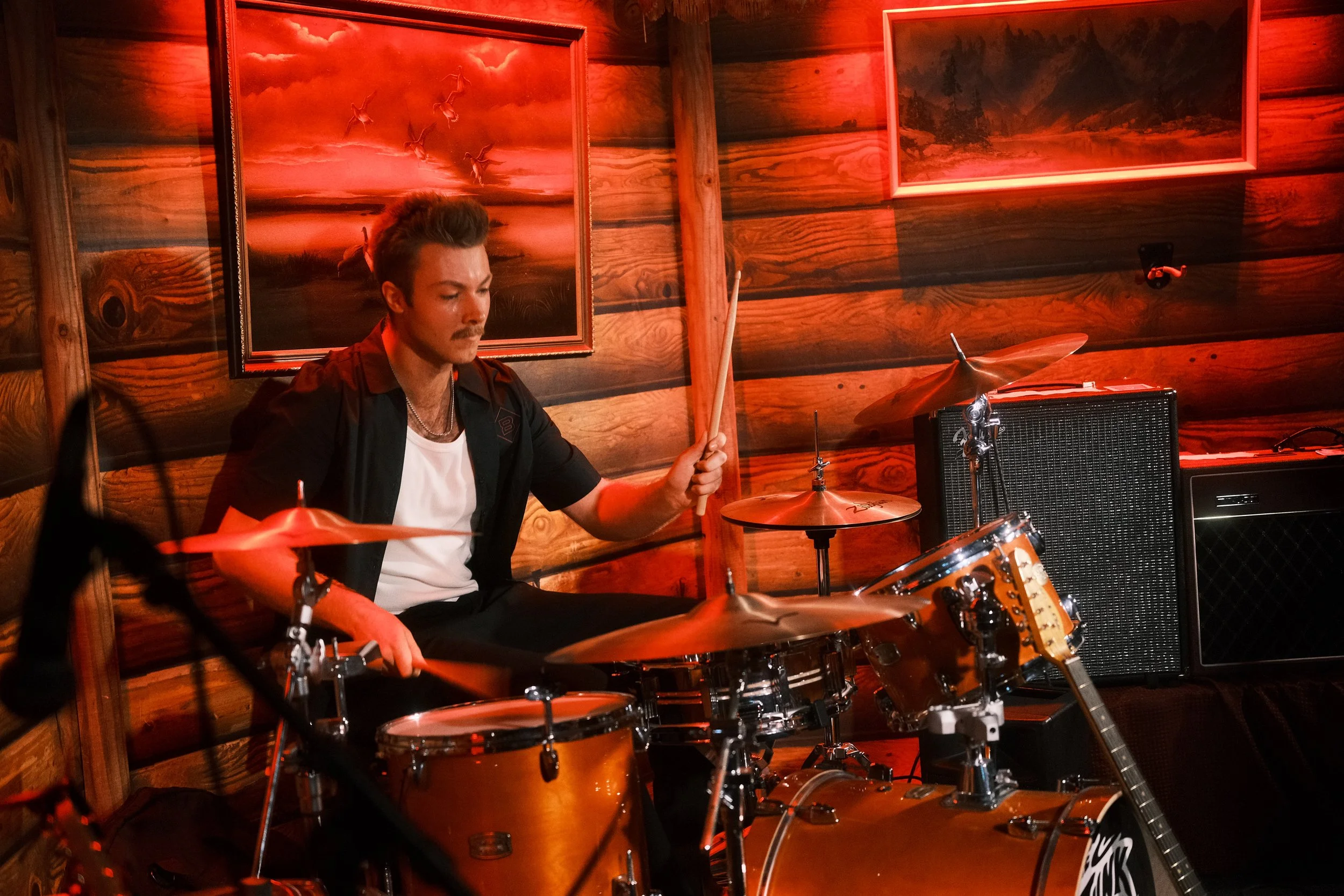 A man playing drums in a wood-paneled room with landscape paintings on the wall.