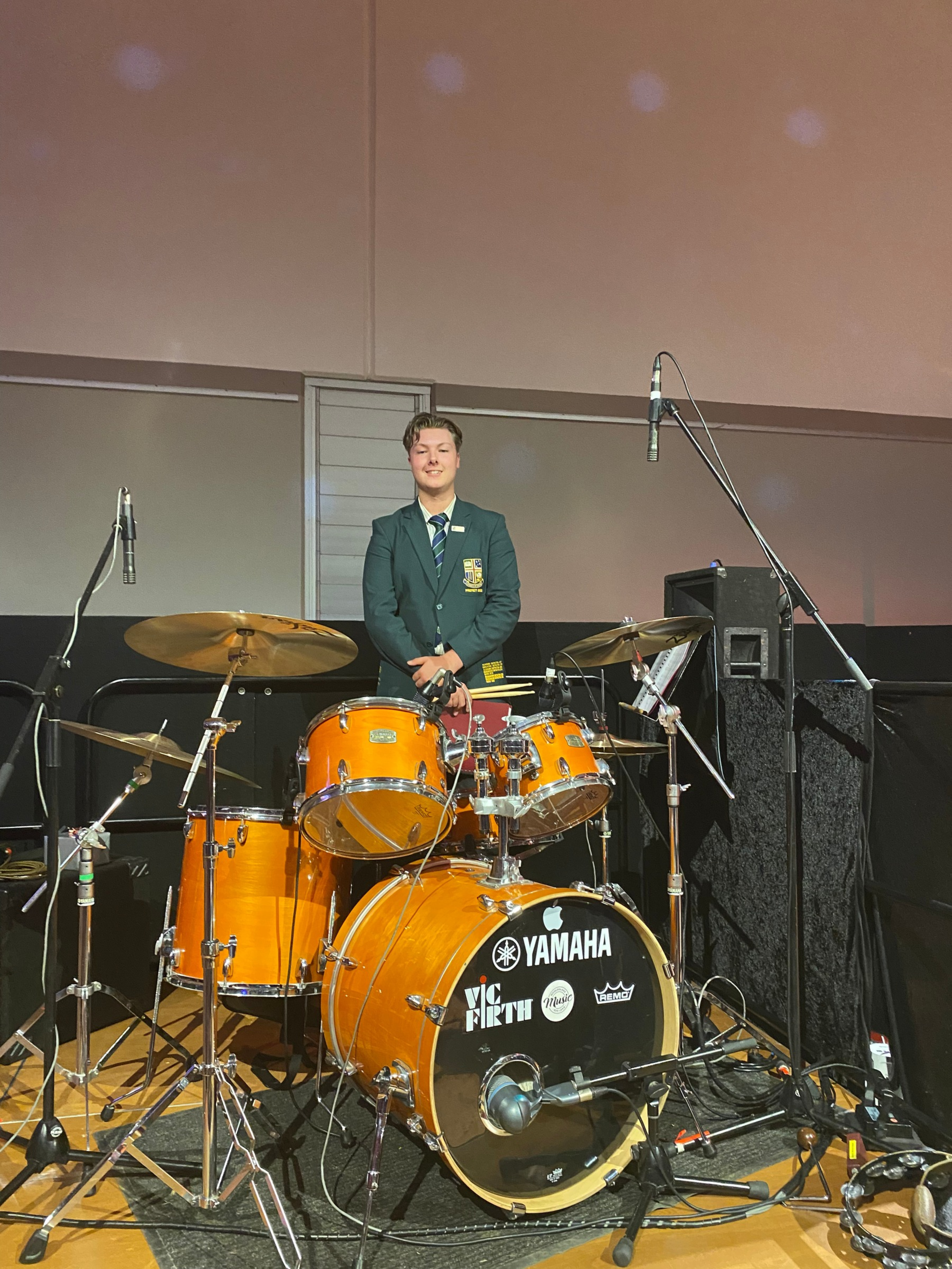 Young person in a dark green blazer and striped tie standing behind a bright orange Yamaha drum set on a stage or in a music studio, with microphones and audio equipment around.