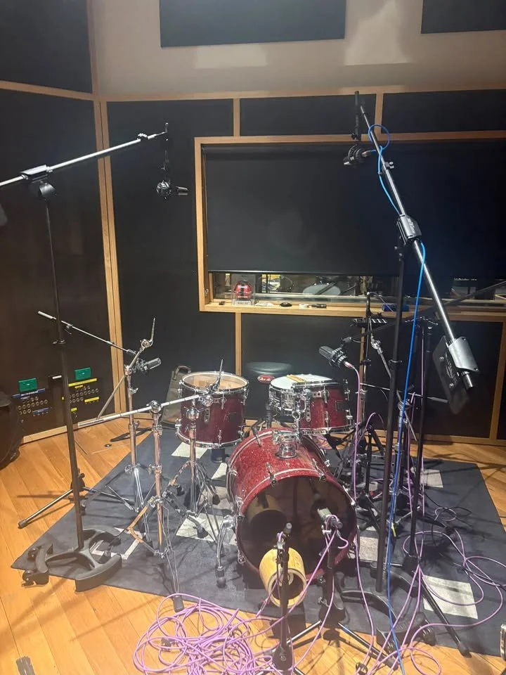 A red drum set in a recording studio, surrounded by several microphones and cables. The studio has wooden floors and soundproof walls with black acoustic panels.