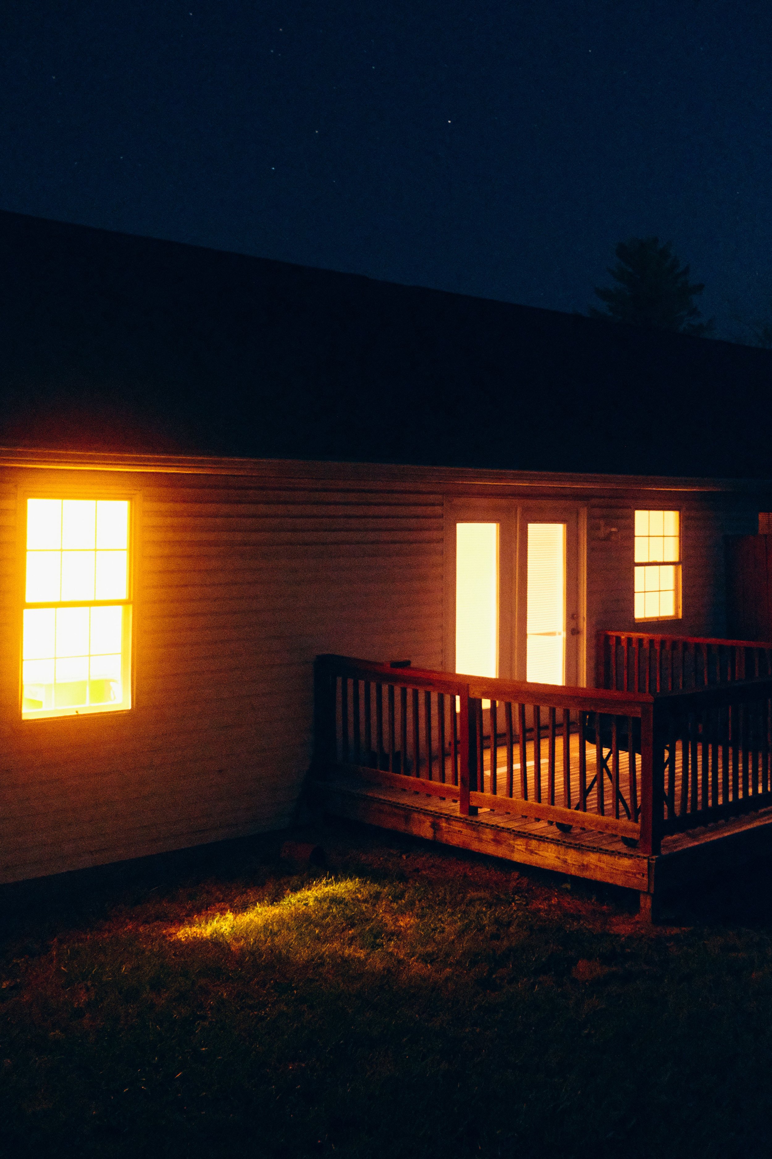 Night scene of a house with illuminated windows, a small wooden porch and railing, surrounded by a dark lawn, with a few stars visible in the sky and a silhouette of a tall tree in the background.
