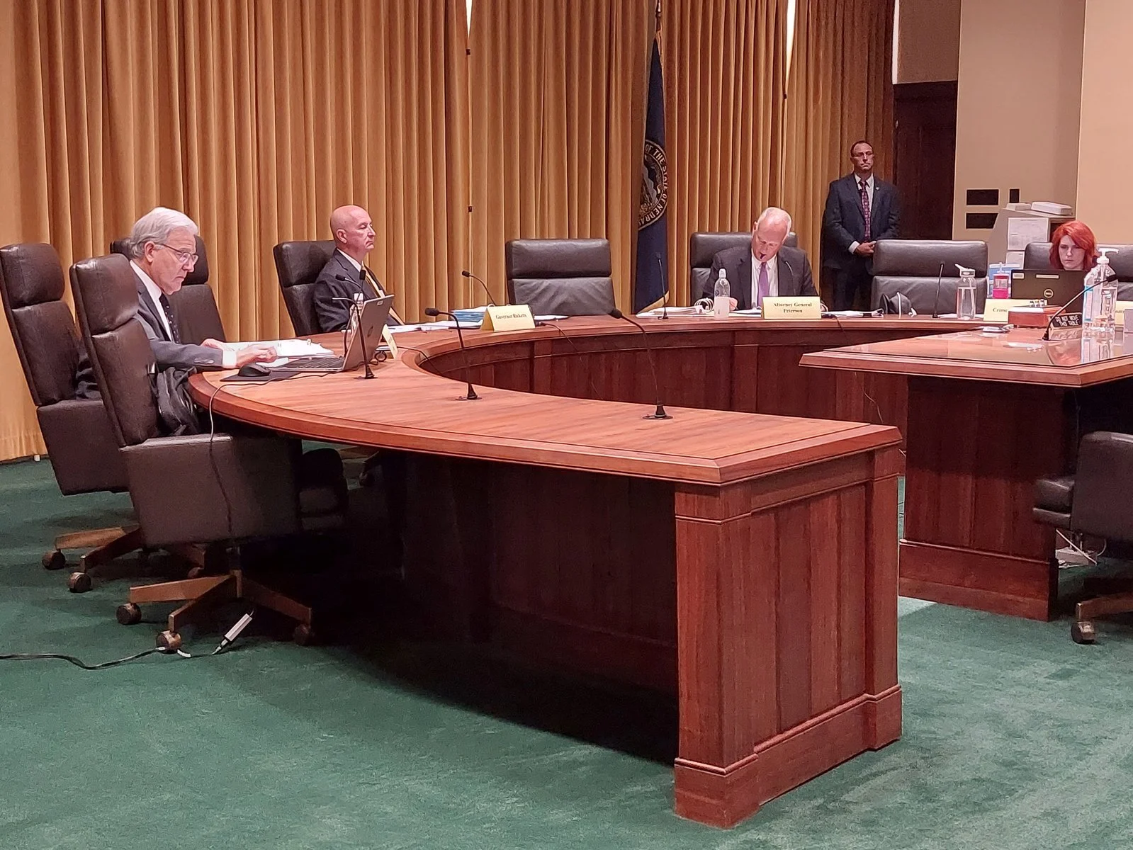 A courtroom with five individuals, three seated at a curved wooden table and two standing. The room has yellow curtains and green carpet. There are nameplates and laptops on the table, with bottles of water and documents. One person has red hair, and one man is wearing a suit and tie.