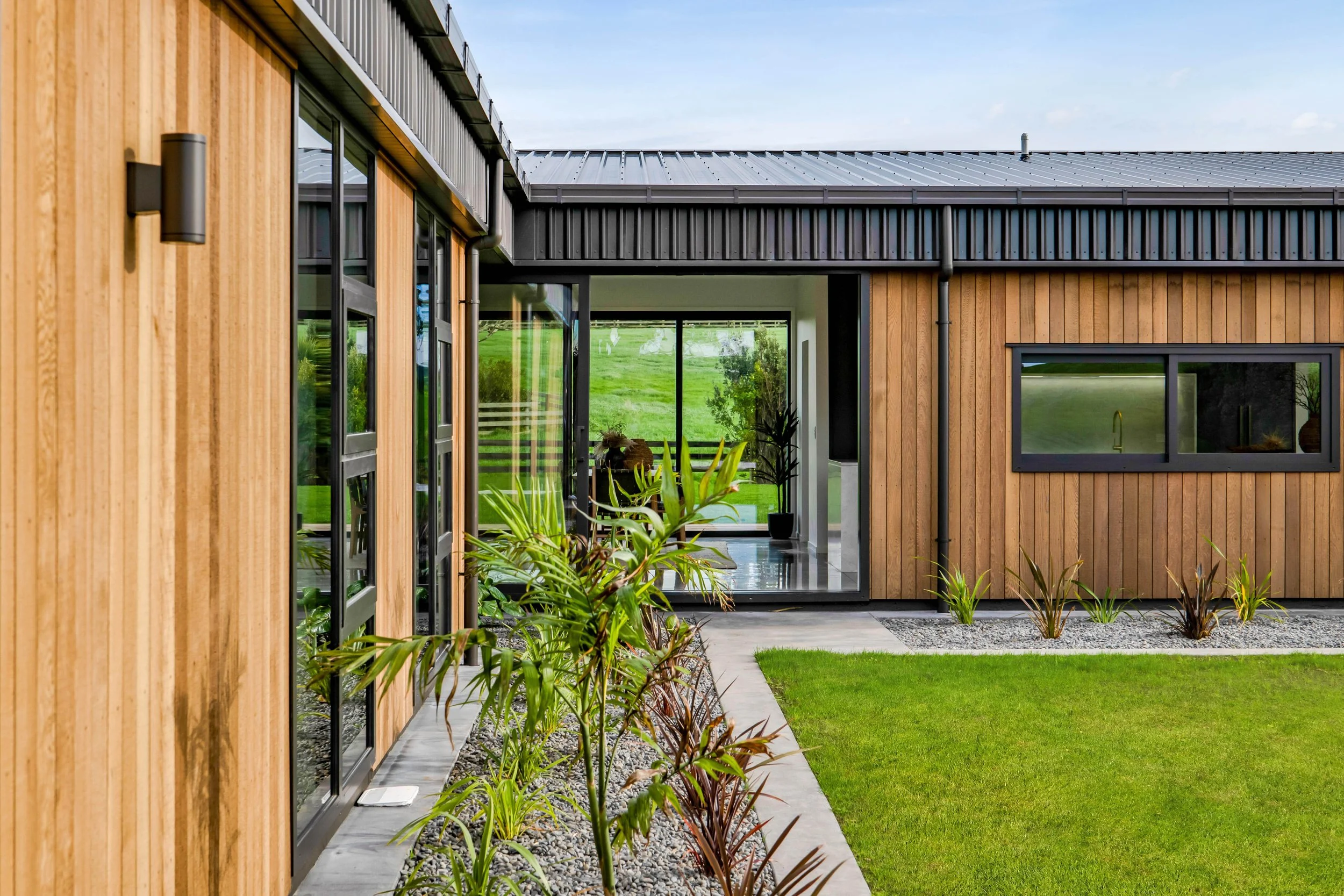 Modern house exterior with wooden siding, large windows, and a landscaped yard with grass and plants. Tapuae subdivision. High quality painting and decorating.