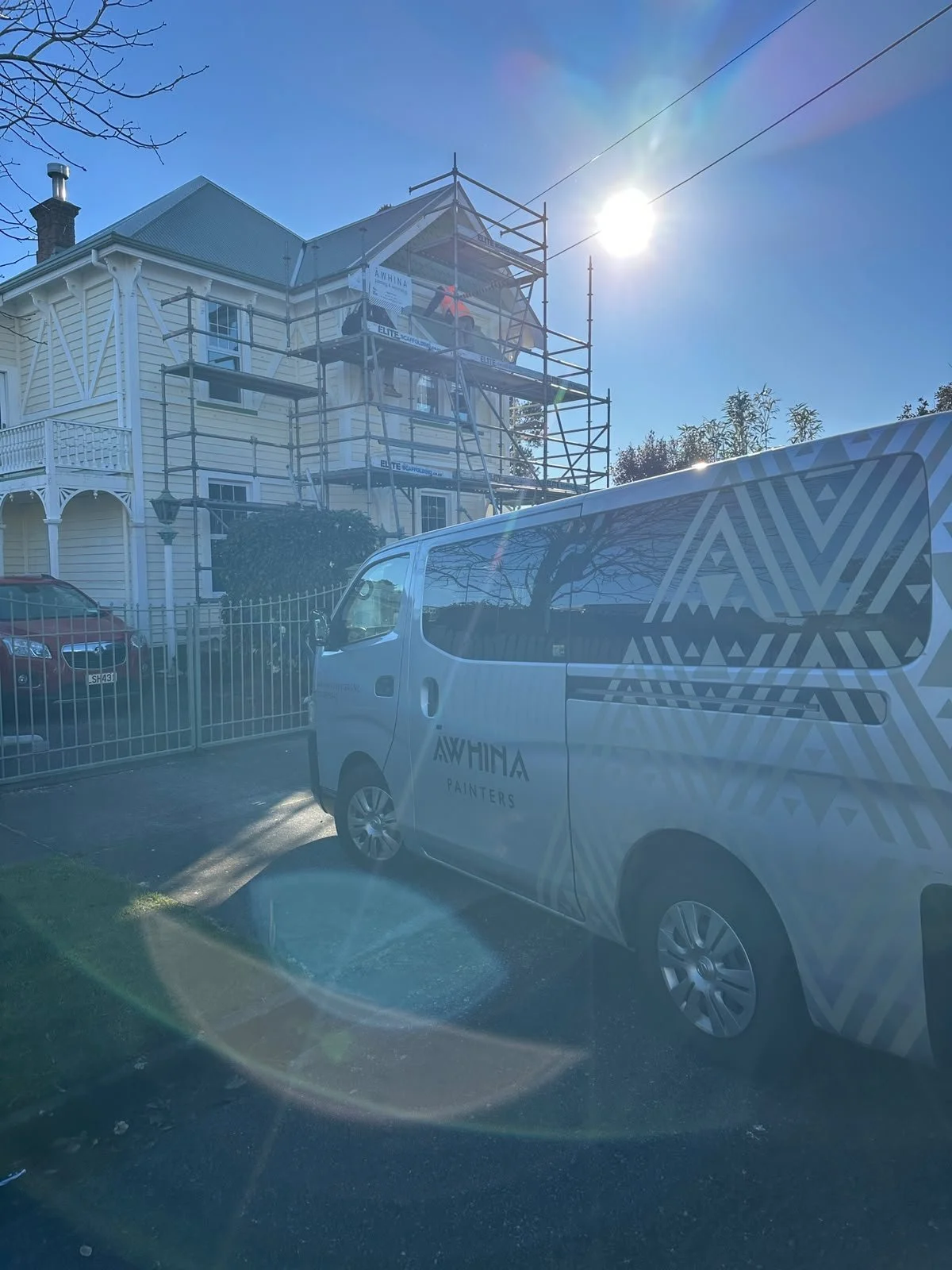 A house under renovation with scaffolding on the front, a white van with 'Awhina Painters' parked outside, and a red car. The sun is shining brightly in the clear blue sky. Dulux accredited painters.