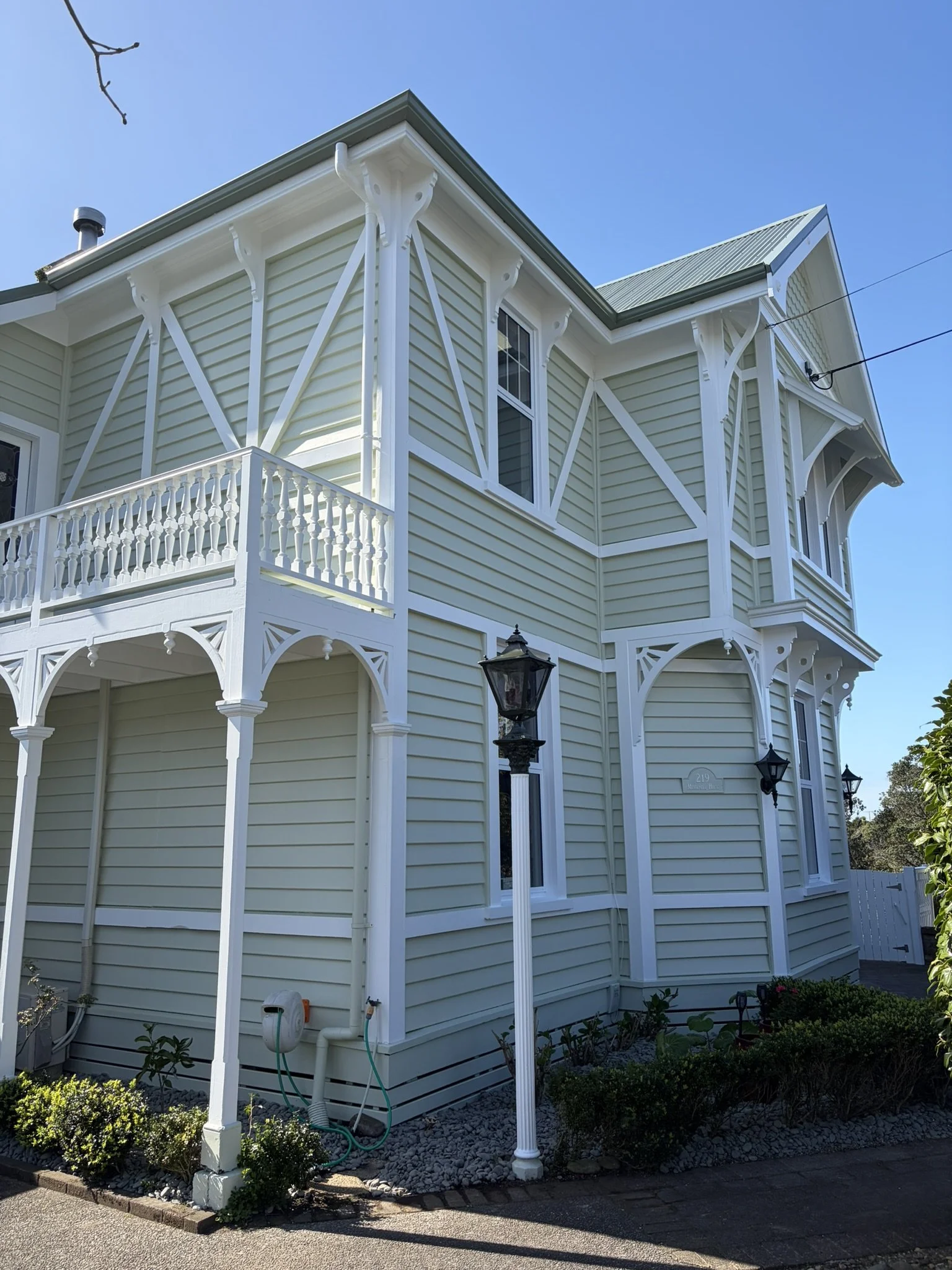Close-up of a light green wooden house with white decorative brackets and trim, showing multiple windows and a balcony with a railing, partly obscured by greenery and a streetlamp. Messenger House built in 1903. Historical building in New Plymouth.