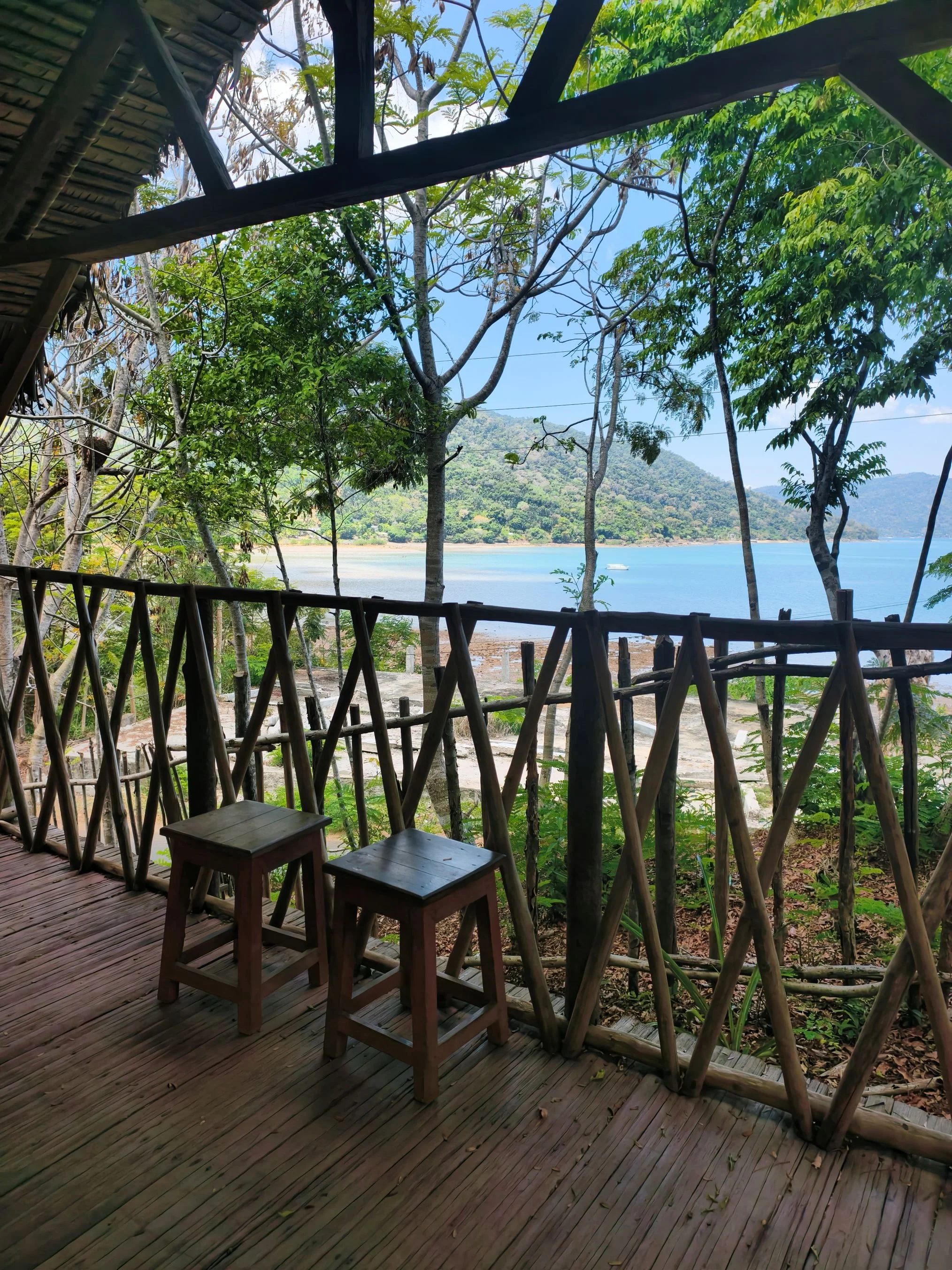 A view from a rustic wooden porch overlooking a tropical lake with green hills in the background, framed by trees and bamboo fencing.