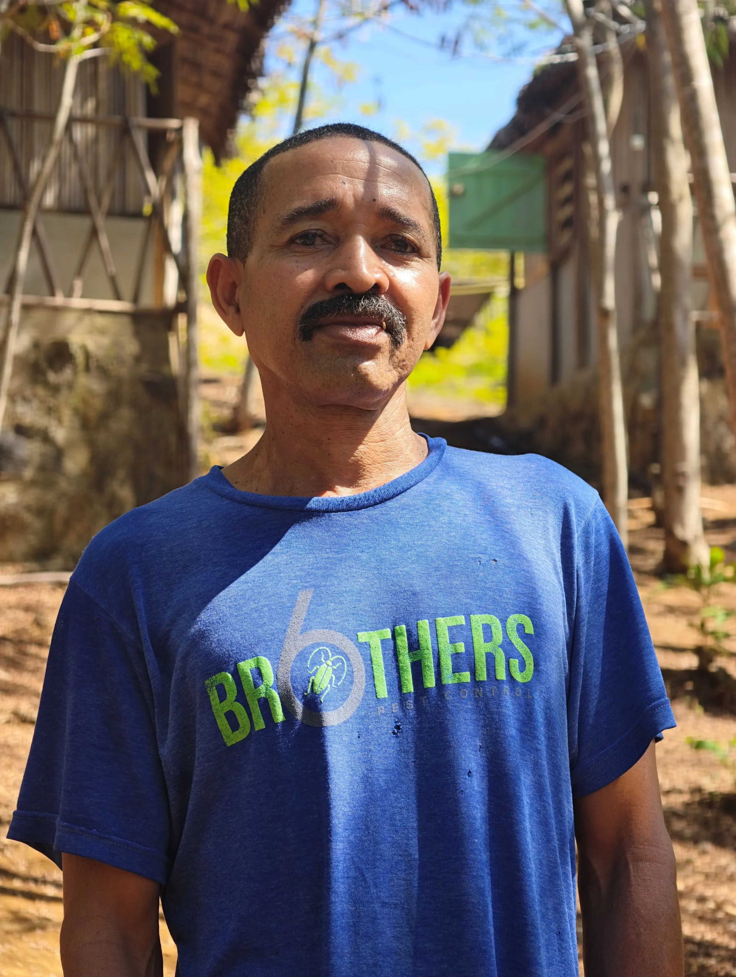 Close-up of a man wearing a blue T-shirt with green and white text, standing outdoors with trees and wooden structures in the background.