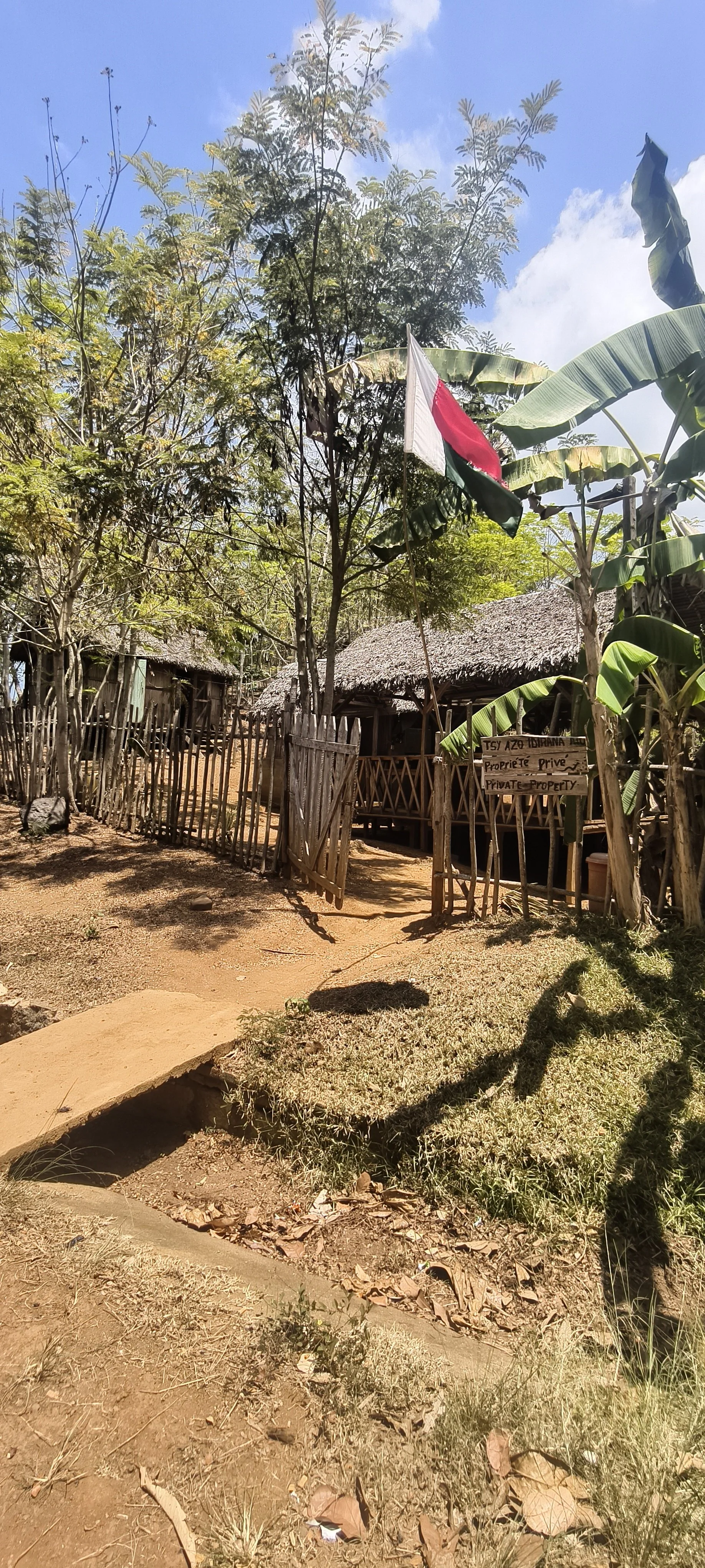 Rural scene with traditional huts made of wood and thatch, surrounded by lush trees and plants, under a blue sky with some clouds. There is a wooden fence, a flag on a pole, and a sign indicating private property.