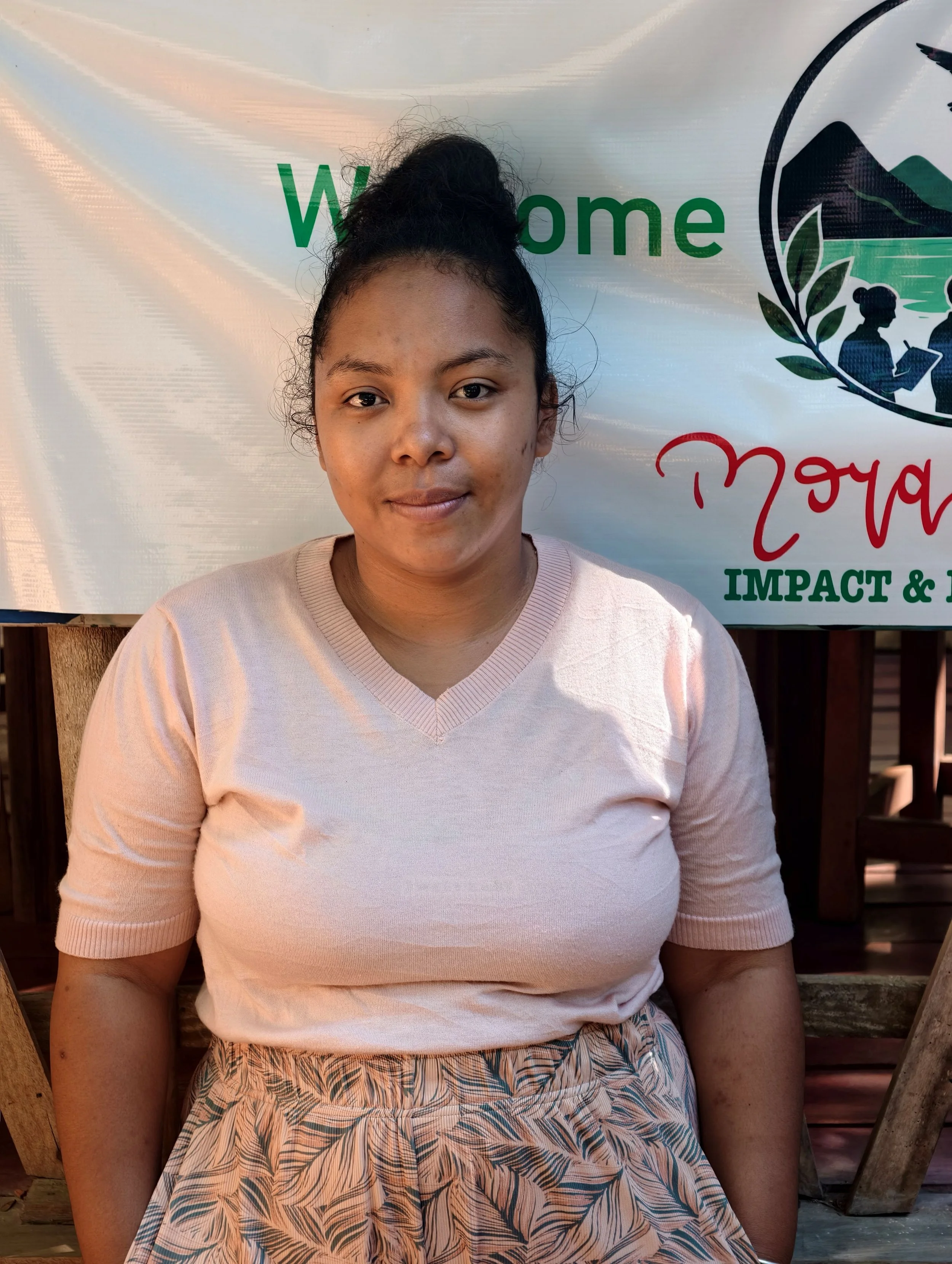 A woman with a high bun hairstyle sitting outside in front of a banner with the word 'Welcome' and a logo that includes mountains and people reading.