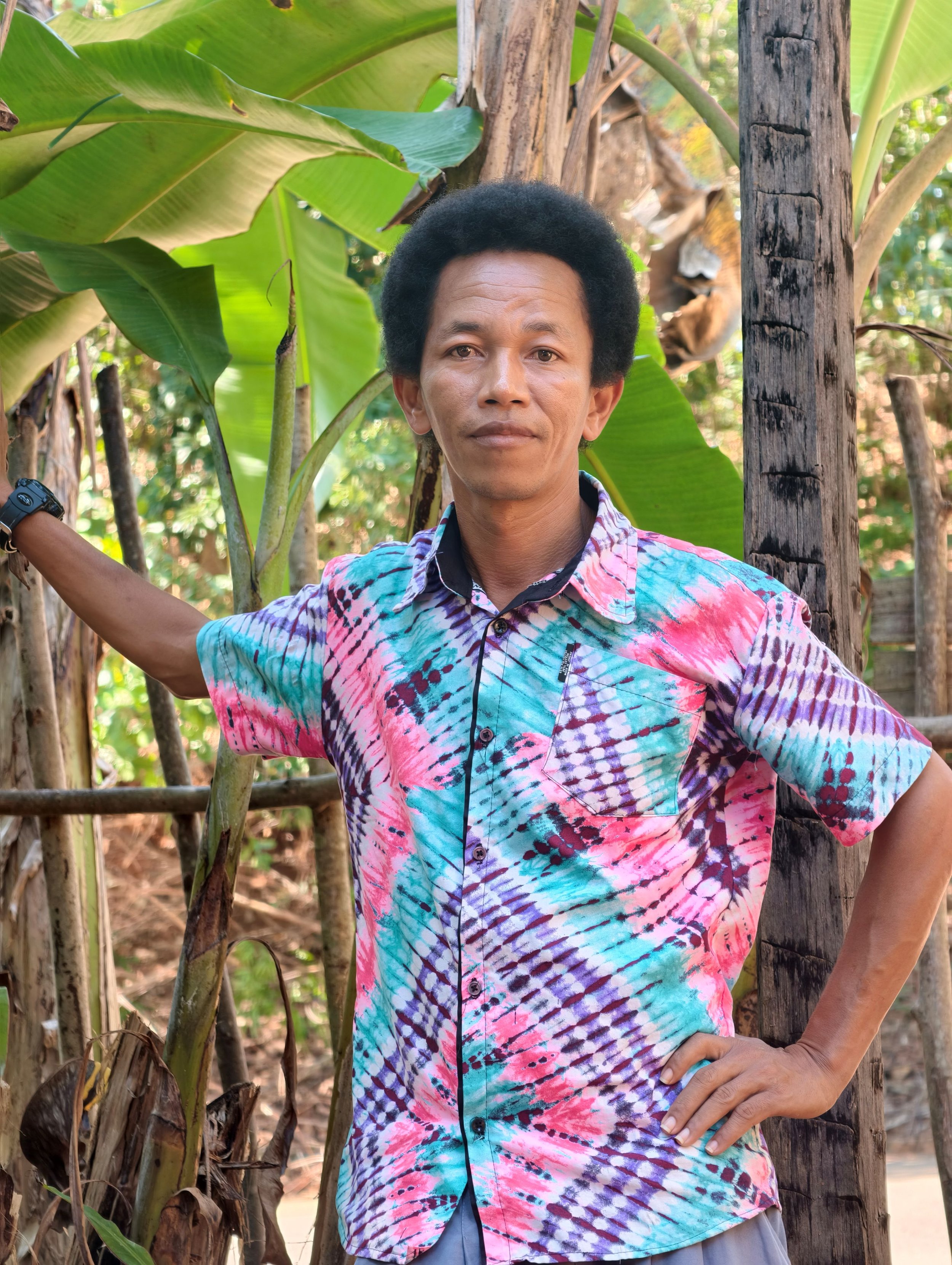 A man standing outdoors among banana plants, wearing a colorful tie-dye shirt with a serious expression, with one hand on his hip and the other resting on a wooden post.