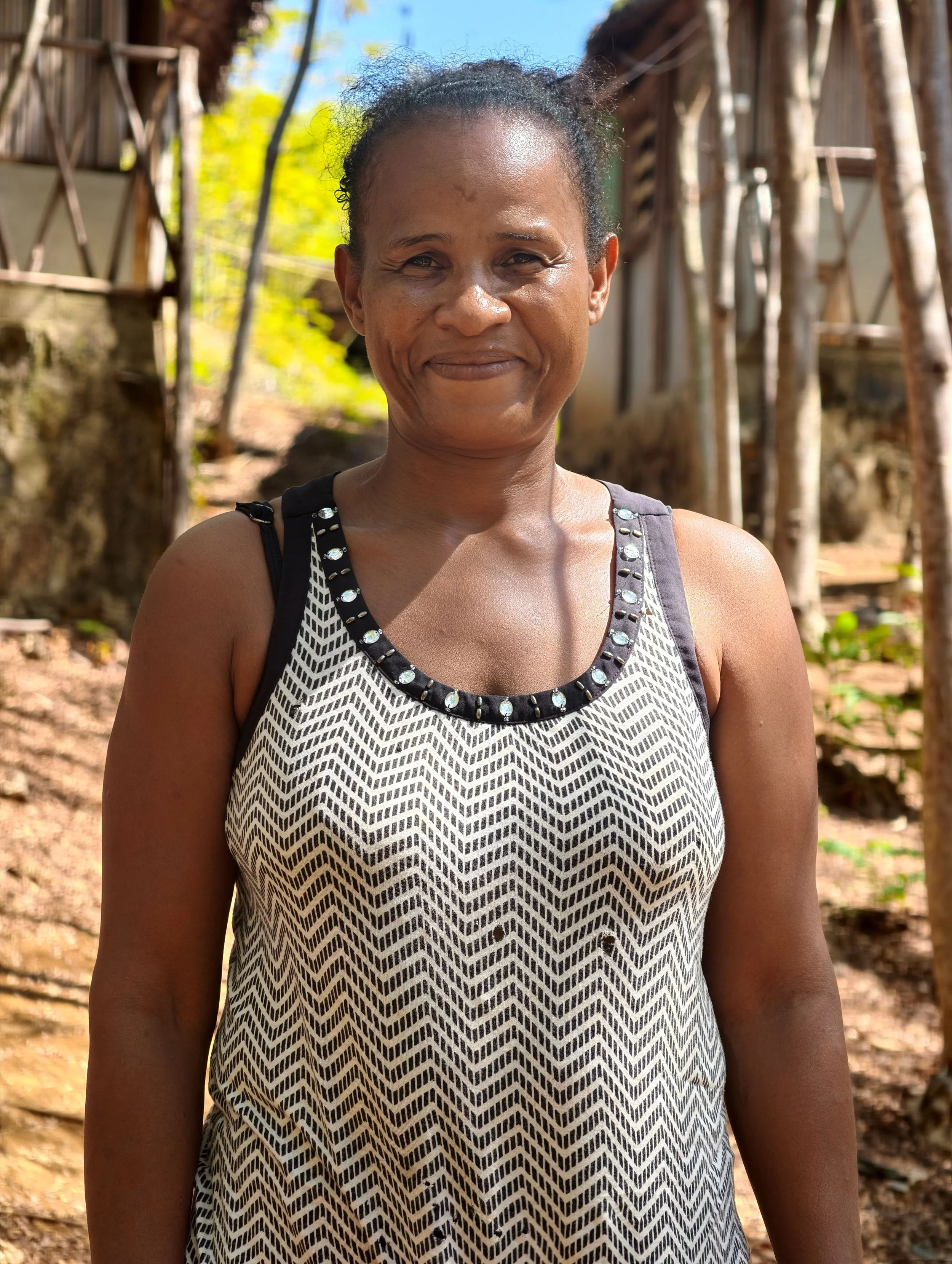 Close-up of a middle-aged woman outdoors smiling, with trees and a wooden structure in the background.