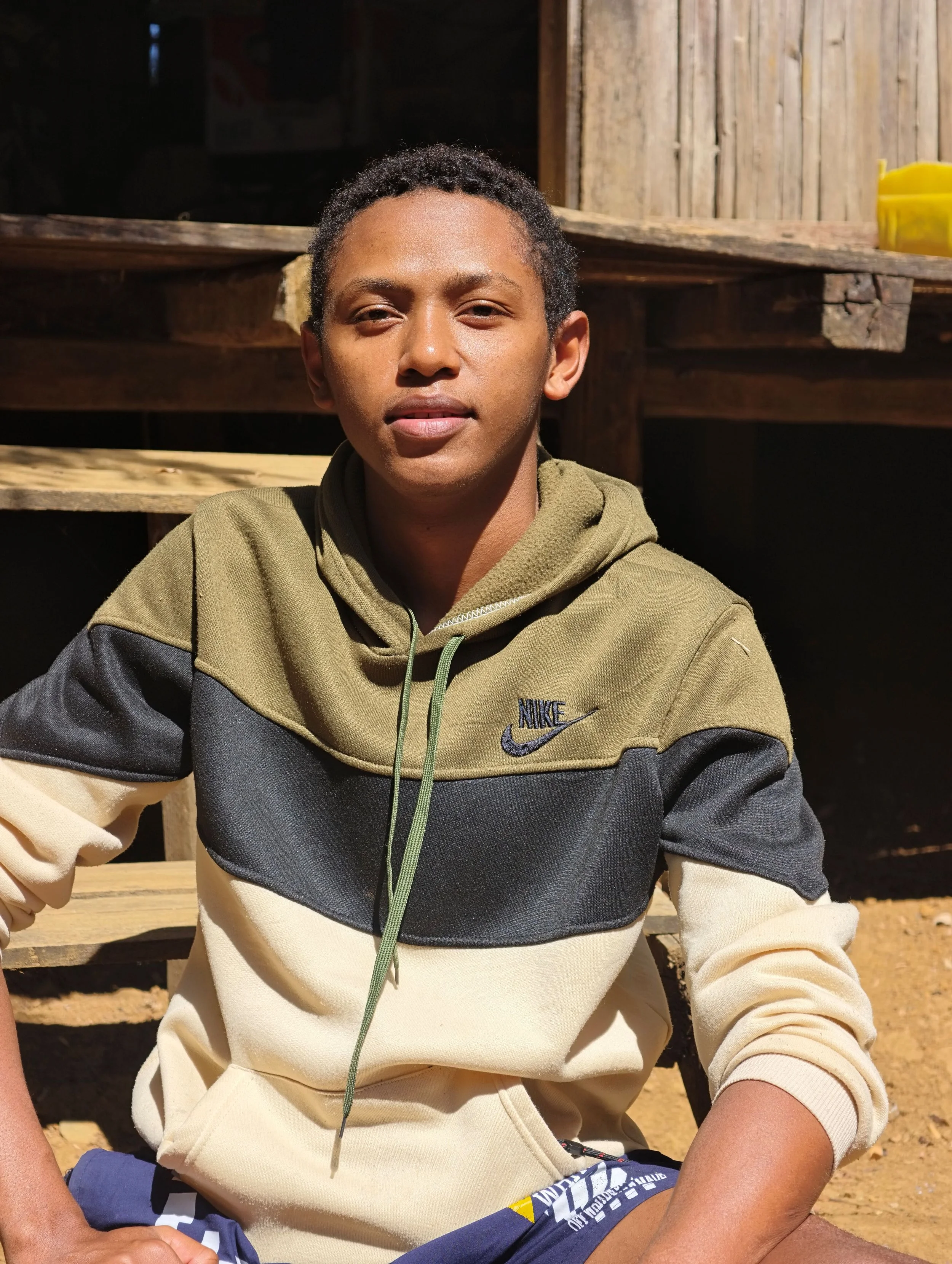A young man with short, curly black hair sitting outdoors. He is wearing a beige and black Nike hoodie and dark shorts, with sunlight illuminating his face. There is a wooden structure behind him and a yellow object on the right side of the background.