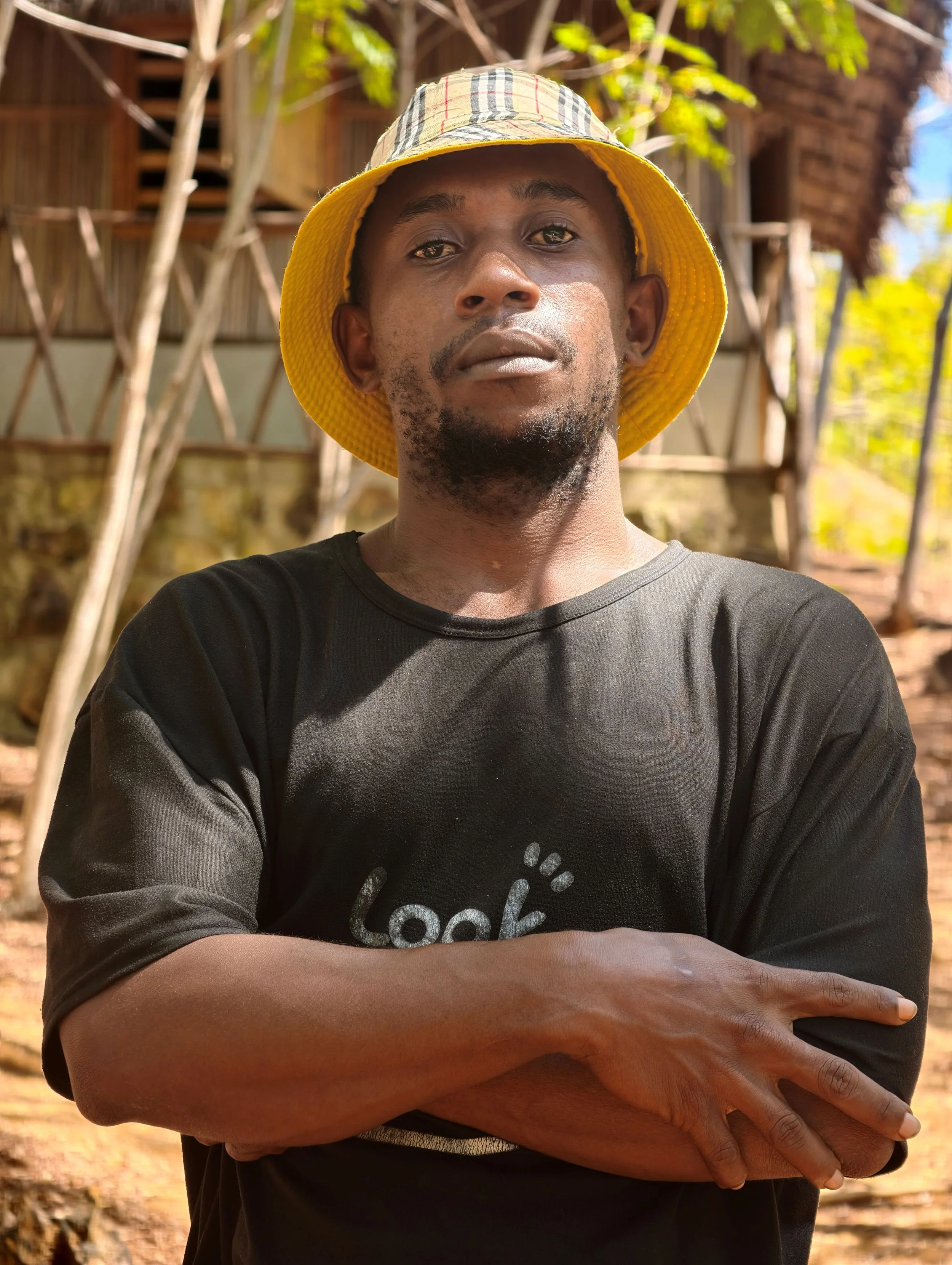 A man wearing a yellow hat and a black t-shirt with a partially visible logo, with his arms crossed, outdoors with some trees and wooden structures in the background.