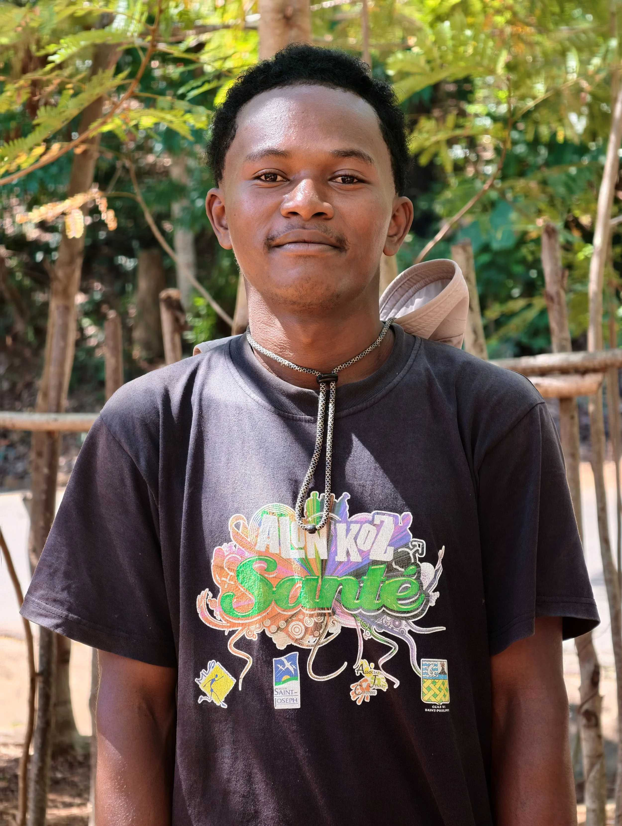 A young man standing outdoors in front of trees and greenery, wearing a black T-shirt with a colorful graphic design and patches, and a necklace.