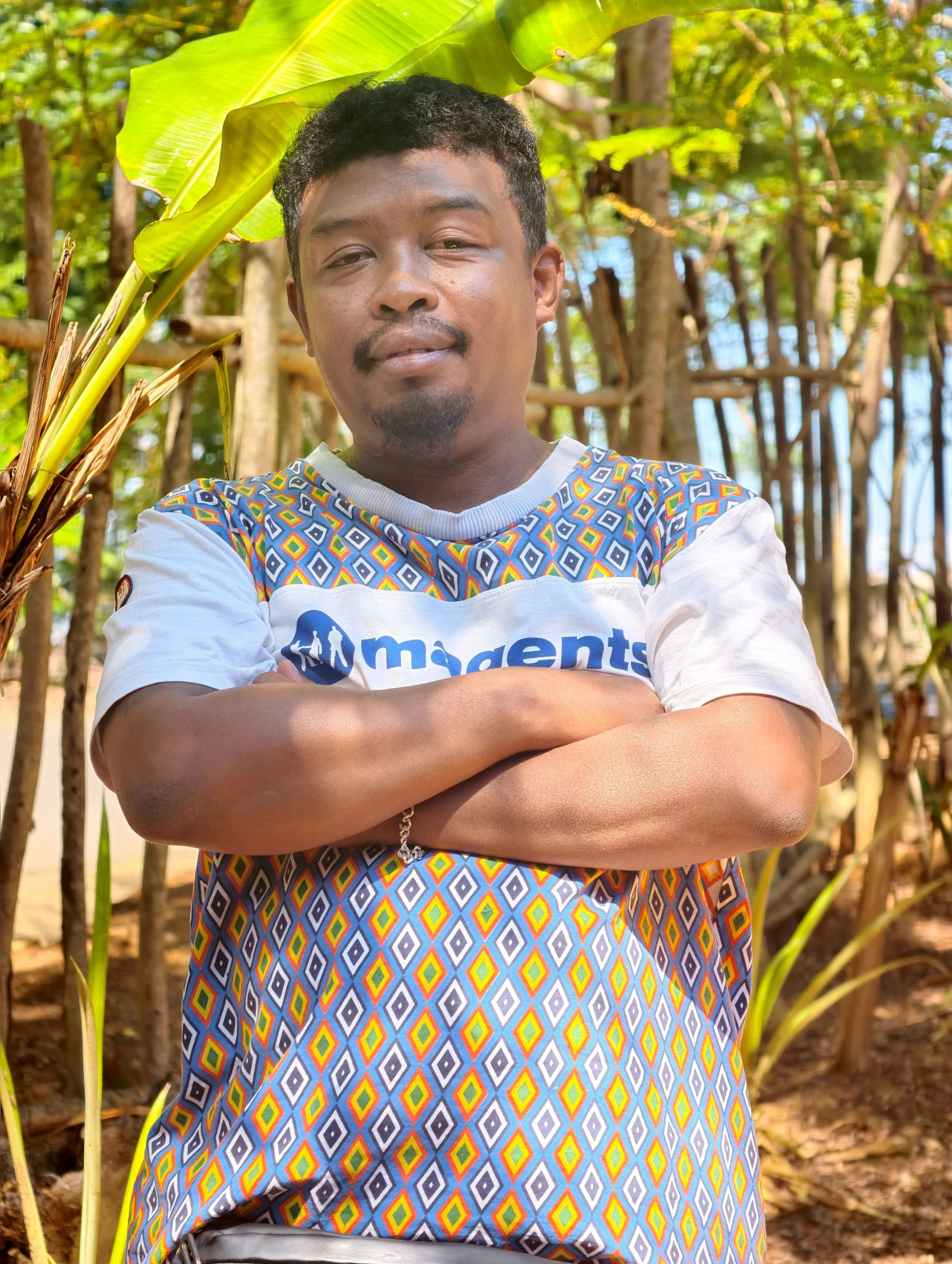 A man standing outdoors with arms crossed in front of lush green trees and plants, wearing a colorful patterned shirt and a white garment with a logo, during daytime.