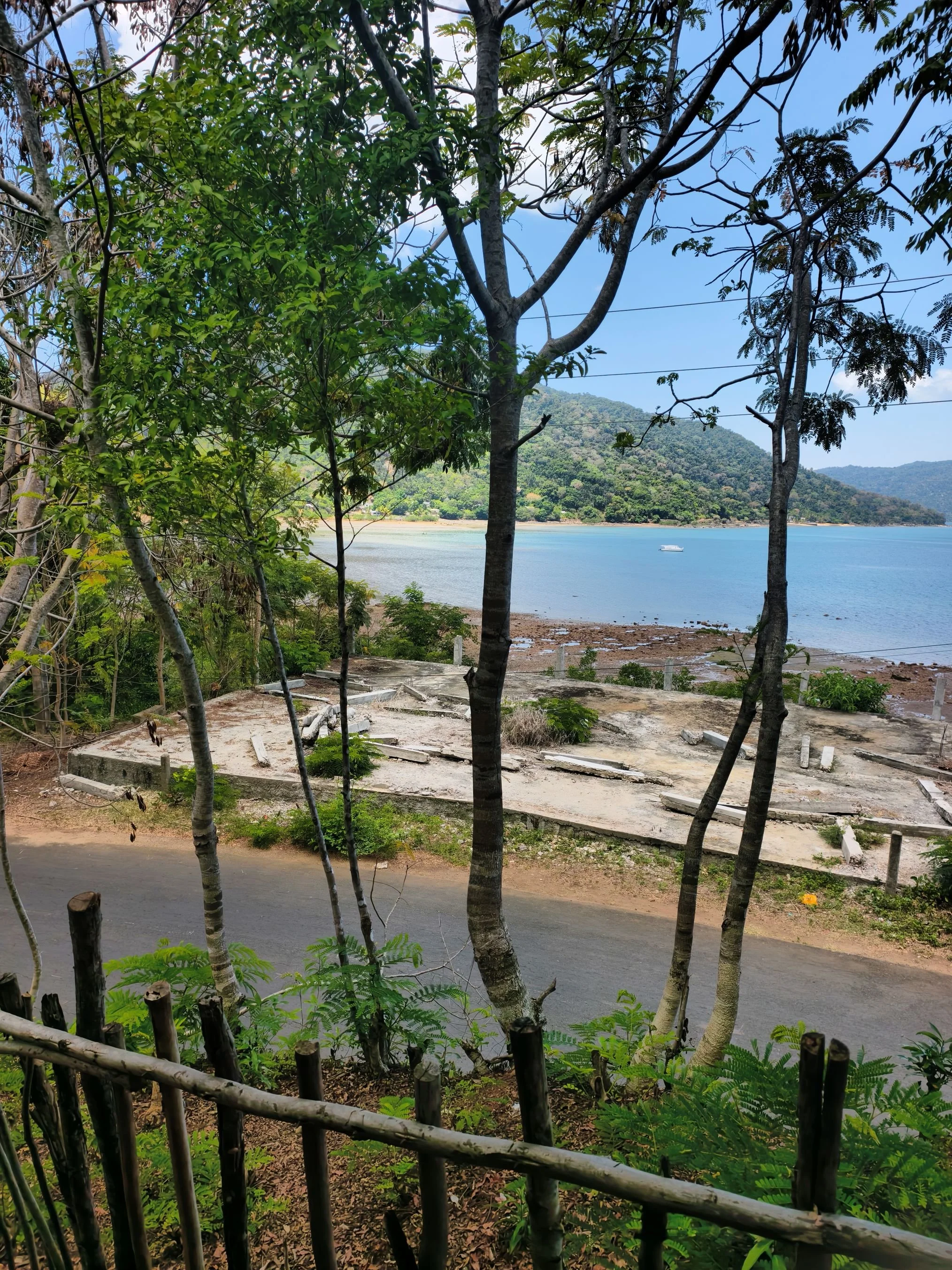 A landscape view of a coastal area with trees in the foreground, a body of water, and hills in the background. There is a boat on the water and some dry land along the shoreline. An unfinished structure is visible in the lower middle part of the imag