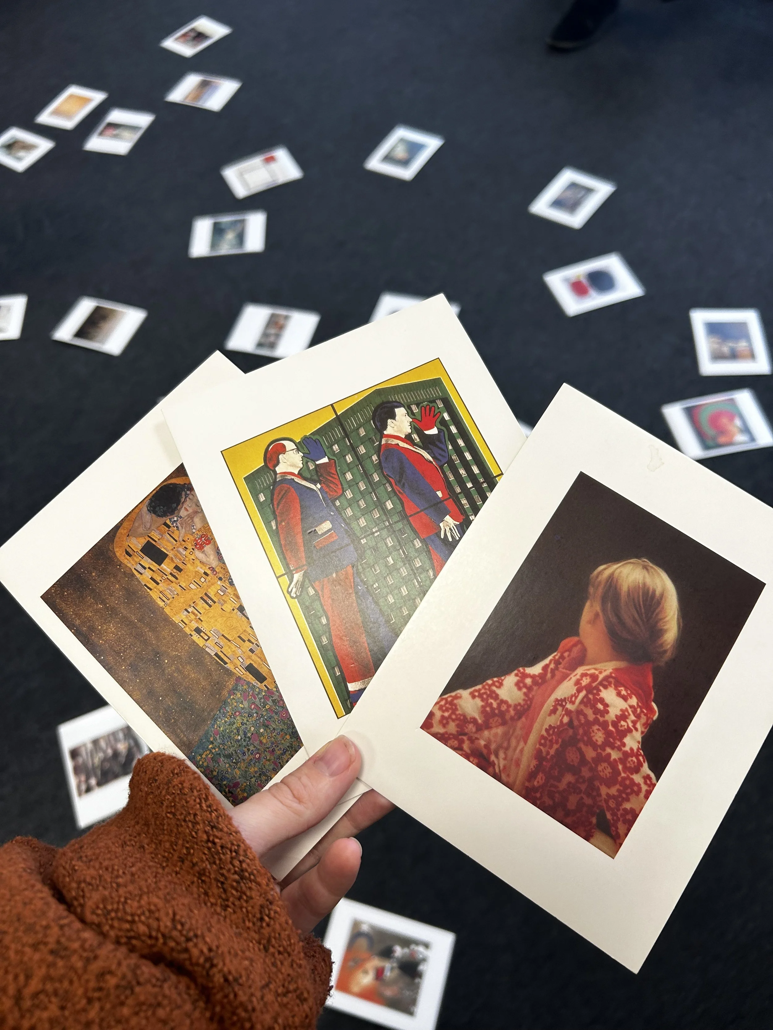A person holding three photographs, with scattered photographs on a dark surface in the background. The photographs feature various scenes, including a woman in red with blonde hair and artwork.