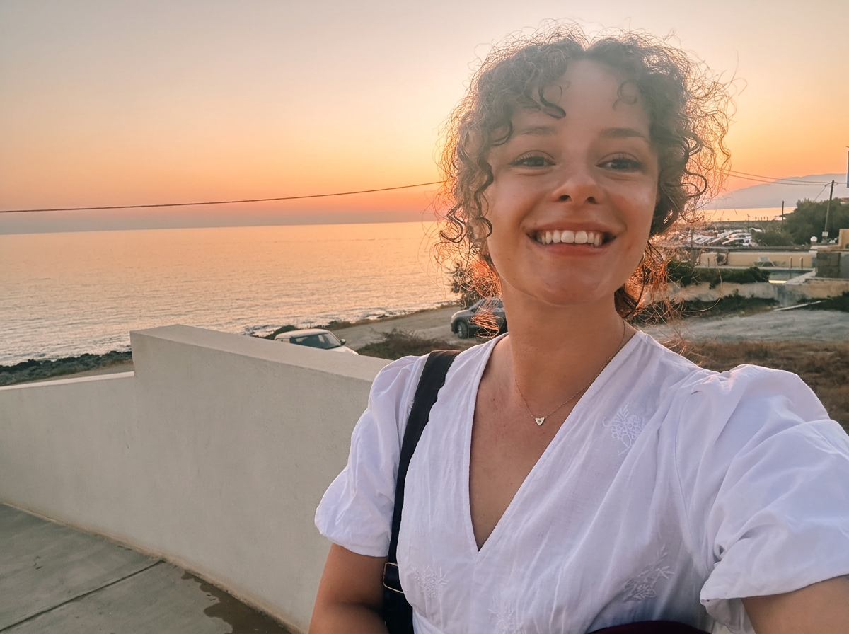 smiling woman with curly hair taking a selfie at sunset near the coast, with a white wall and parked cars in the background