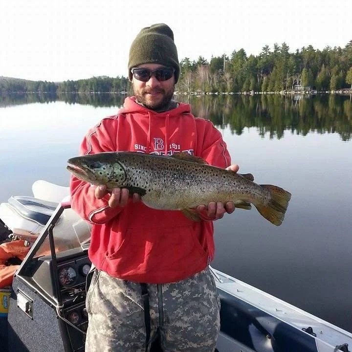 A man in a red hoodie, camouflage pants, sunglasses, and a beanie holding a large fish on a boat by a calm lake with trees in the background.