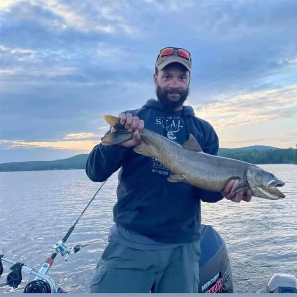 Man holding a large fish on a boat, with a lake and hills in the background, during sunset.