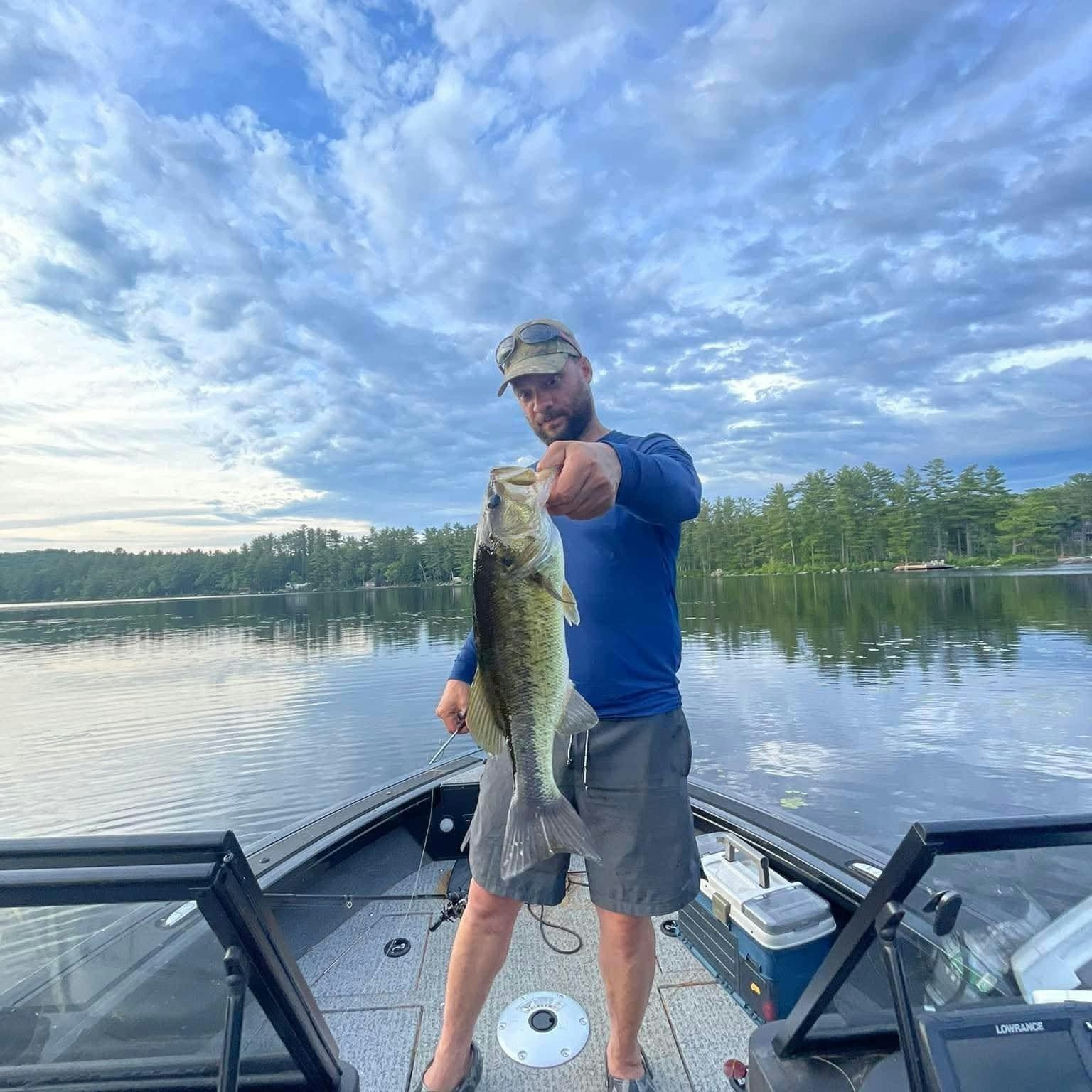 A man on a boat holding a large bass fish he caught, with a lake and trees in the background under a partly cloudy sky.