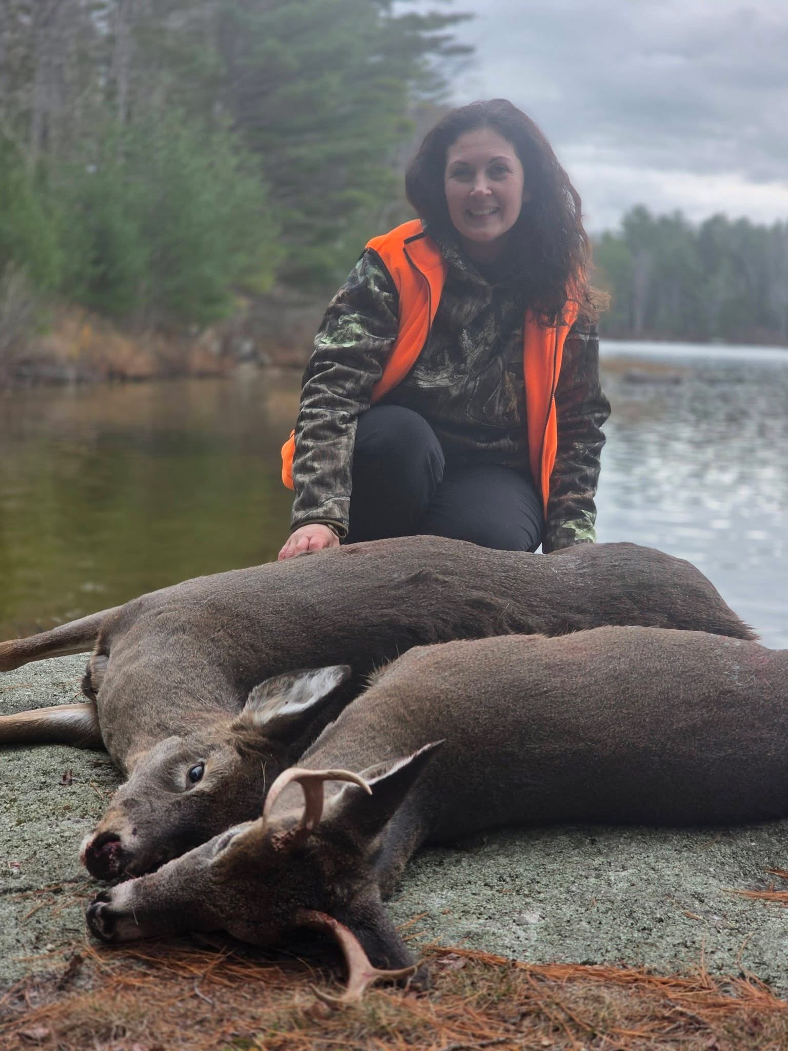 A woman dressed in camouflage and orange outdoor gear kneels by dead animals, possibly deer, lying on the ground near a river with trees and cloudy sky in the background.