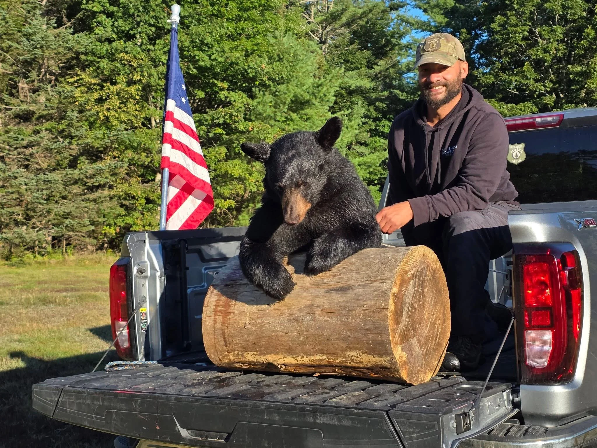 A man with a beard and wearing a camouflage cap with a 'Wilderness Guide' patch, smiling while sitting on the tailgate of a silver pickup truck. In front of him on the truck bed is a large, black bear hide stretched over a wooden frame, and a small American flag is positioned nearby. The background is filled with green trees and blue sky.