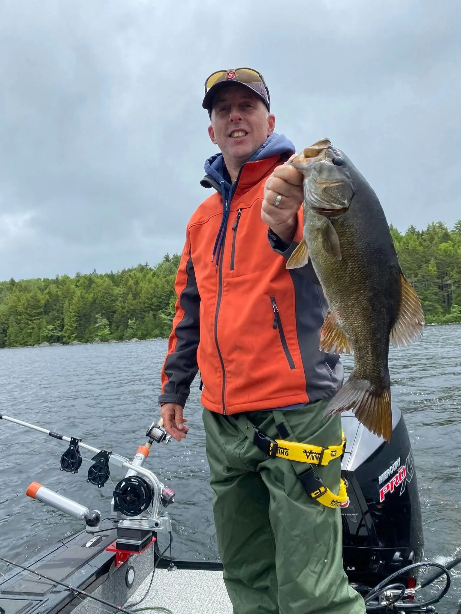 A man holding a large fish on a boat with water and trees in the background.