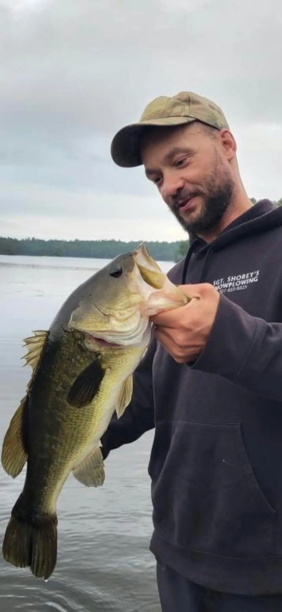 Man holding a large fish near a lake on a cloudy day.