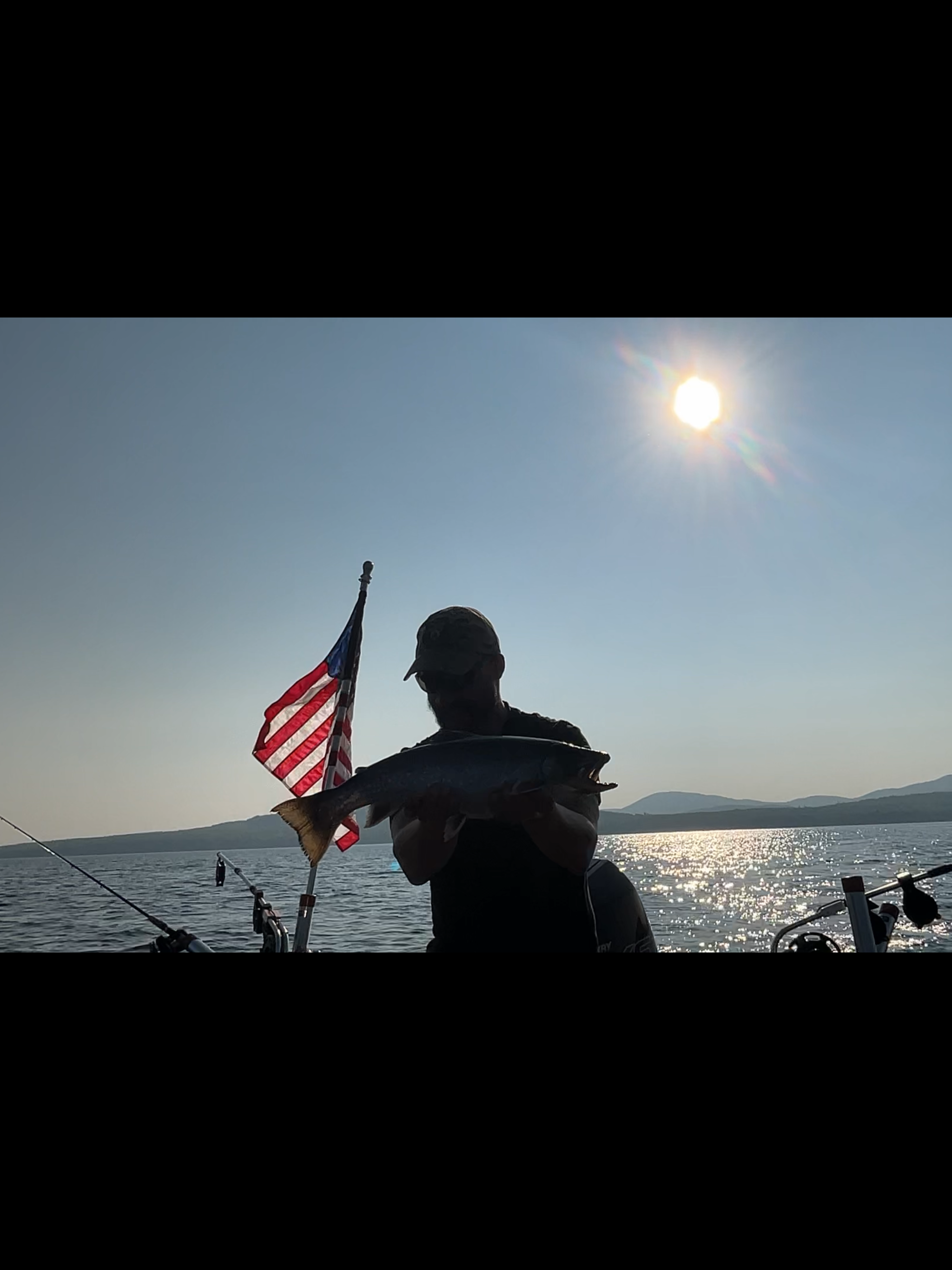 A person holding a fish and an American flag on a boat with a lake and mountains in the background, under a bright sun.