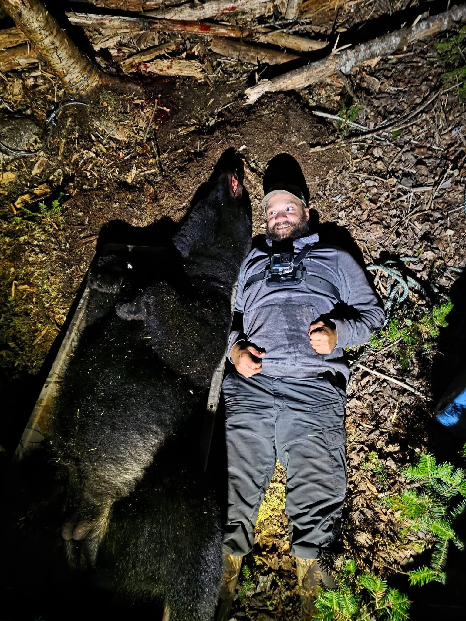 A man lying on the ground in a forest with a large black bear. The man is smiling and looking up at the camera, with a GoPro camera strapped to his chest. The bear is nearby, facing away from the man, and appears to be wet.
