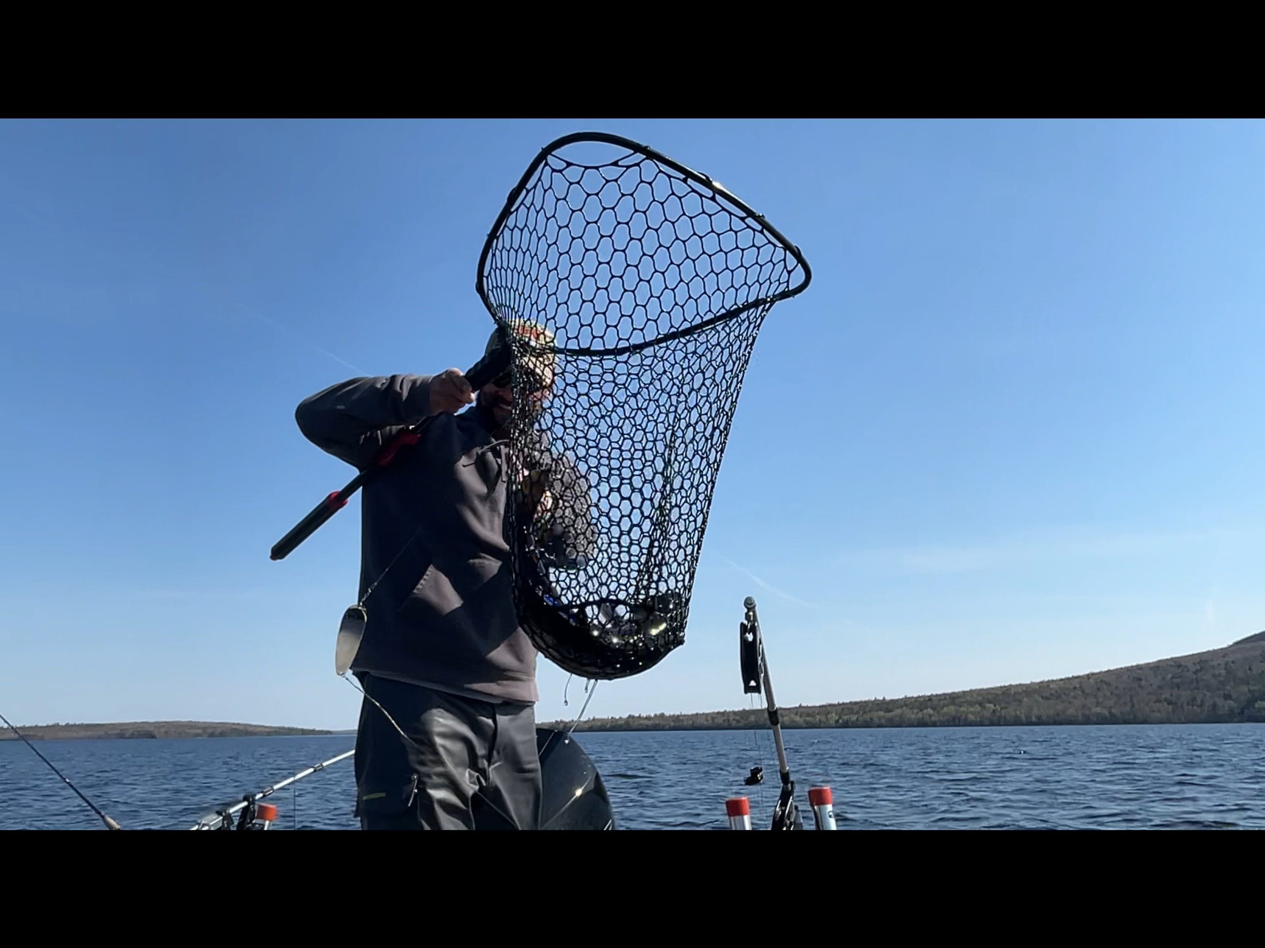 Person fishing on a boat in a lake, holding a fishing net and standing near fishing equipment with hills in the background under a clear blue sky.