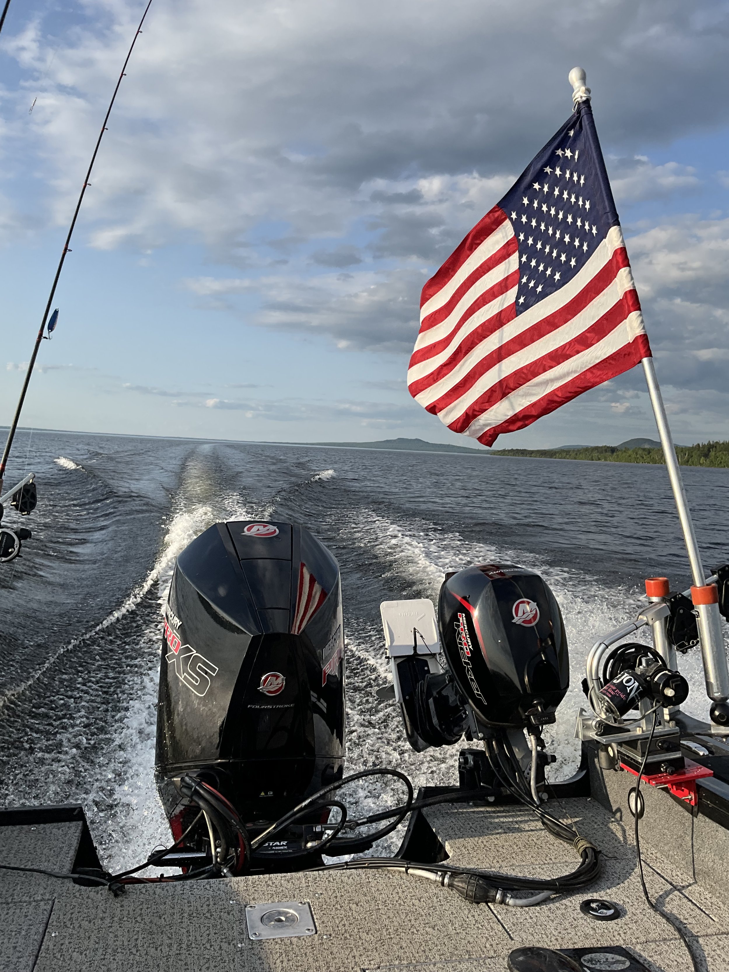 View from the back of a boat showing the water with a wake pattern, two outboard engines, and a mounted motorbike throttle control with American flag fluttering in the wind on a pole. The sky is partly cloudy with distant land visible.