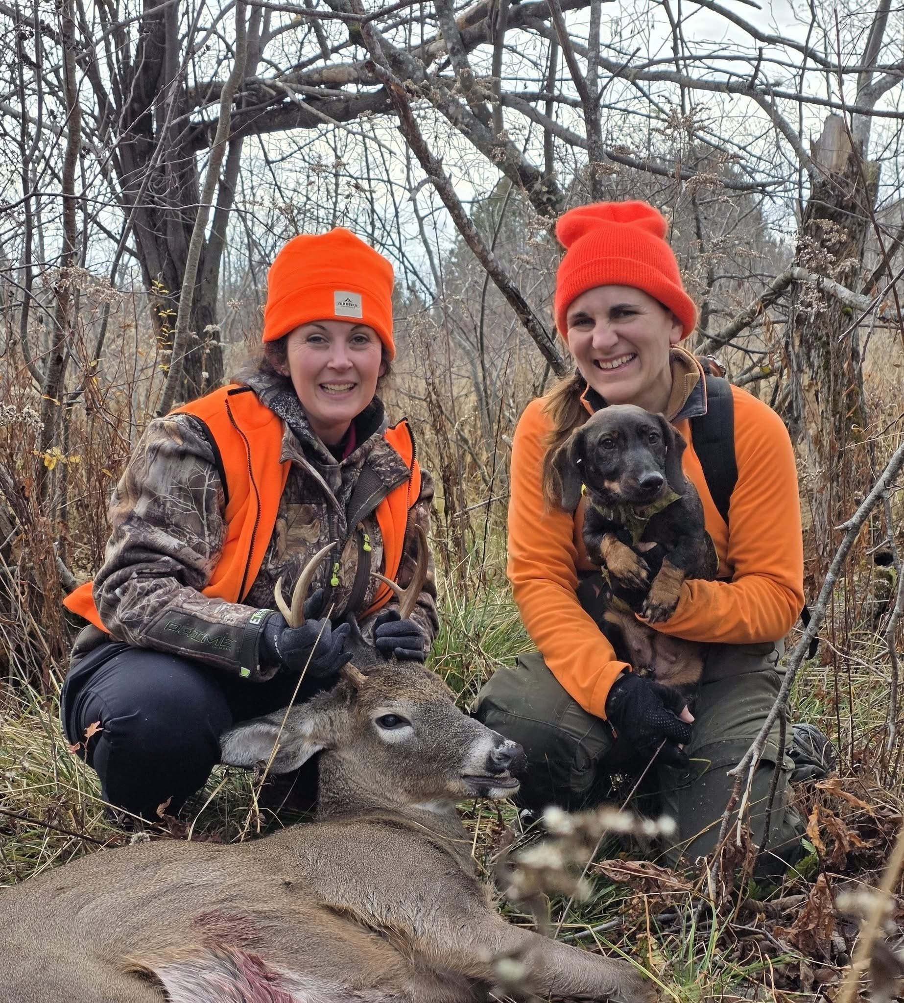 Two women are outdoors in a wooded area, wearing hunting gear and bright orange hats. They are smiling, with one woman holding onto a large, dead deer and a dog. The women's faces and the deer are in focus, with bare trees and branches in the background.