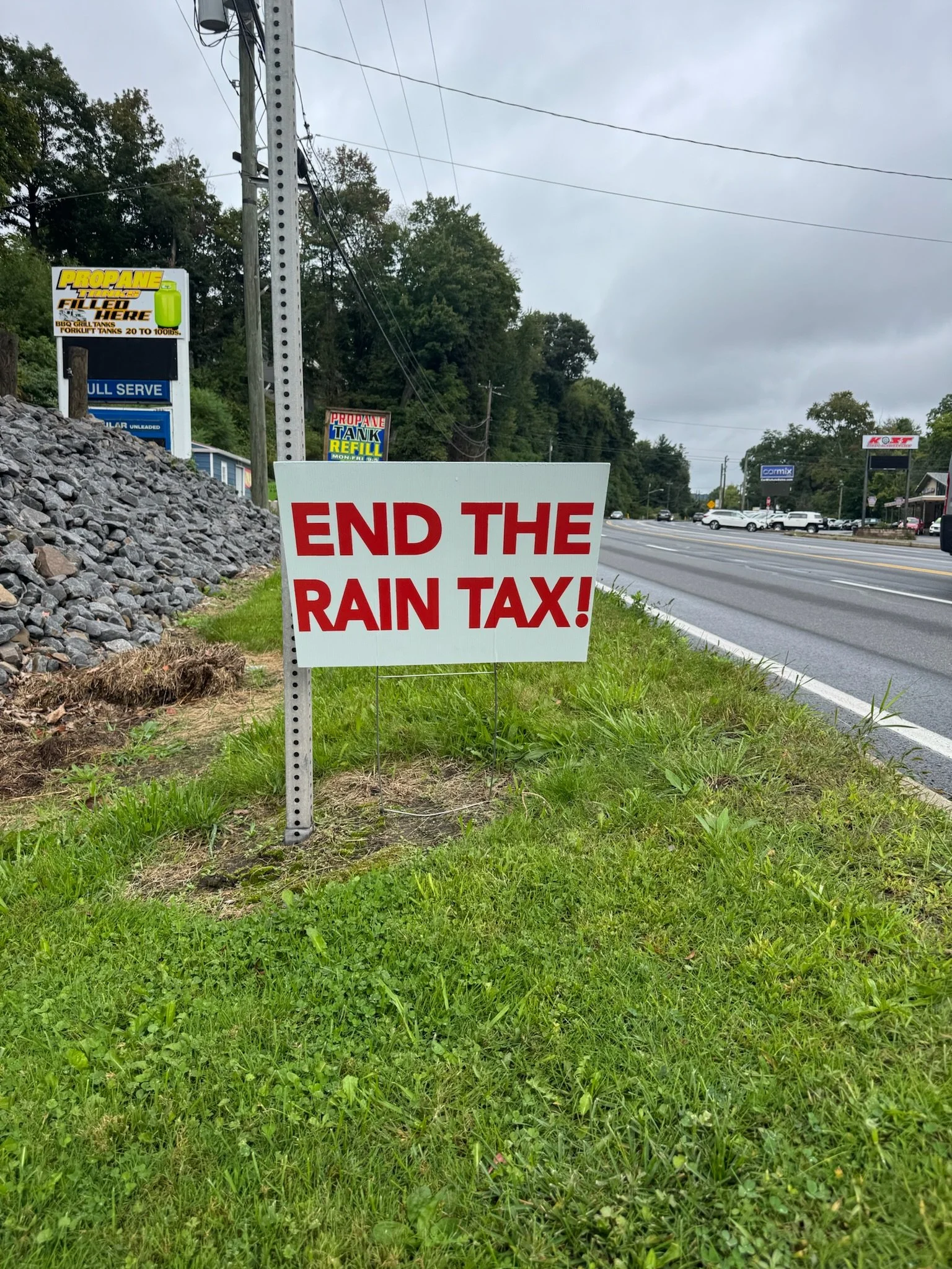 A roadside sign that reads 'END THE RAIN TAX!' in red capital letters on a white background, with a grassy area and commercial buildings in the background.