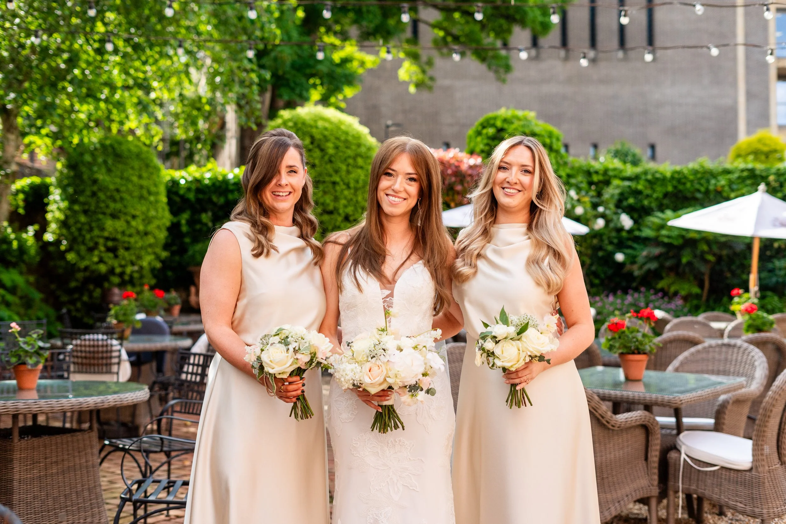 Three women standing together at a wedding reception outdoors, dressed in elegant white dresses, holding bouquets of white and blush roses, smiling with greenery and string lights in the background.