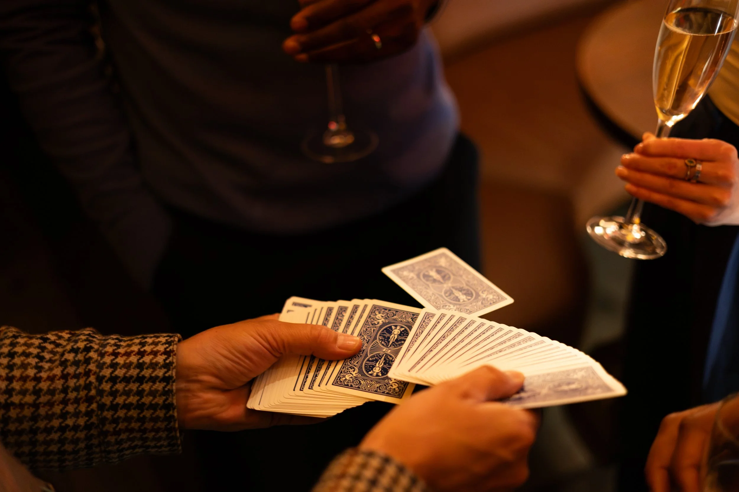 People playing a card game, with one person holding playing cards and another holding a glass of champagne.