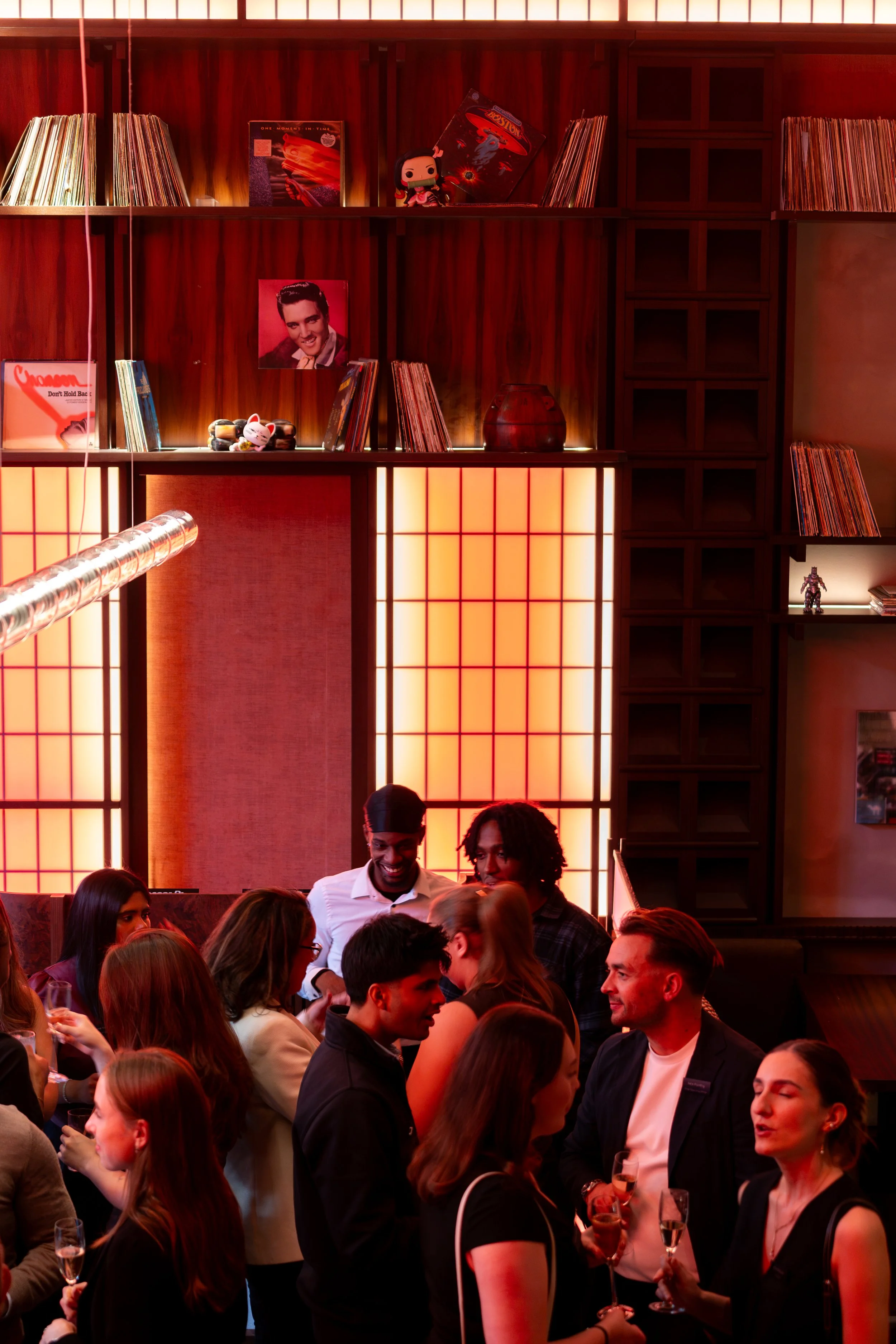 People gathered in a warmly lit restaurant or bar, engaging in conversation and holding drinks. The background features wooden shelving with vinyl records and decorative items, including posters and figurines, illuminated with soft lighting.